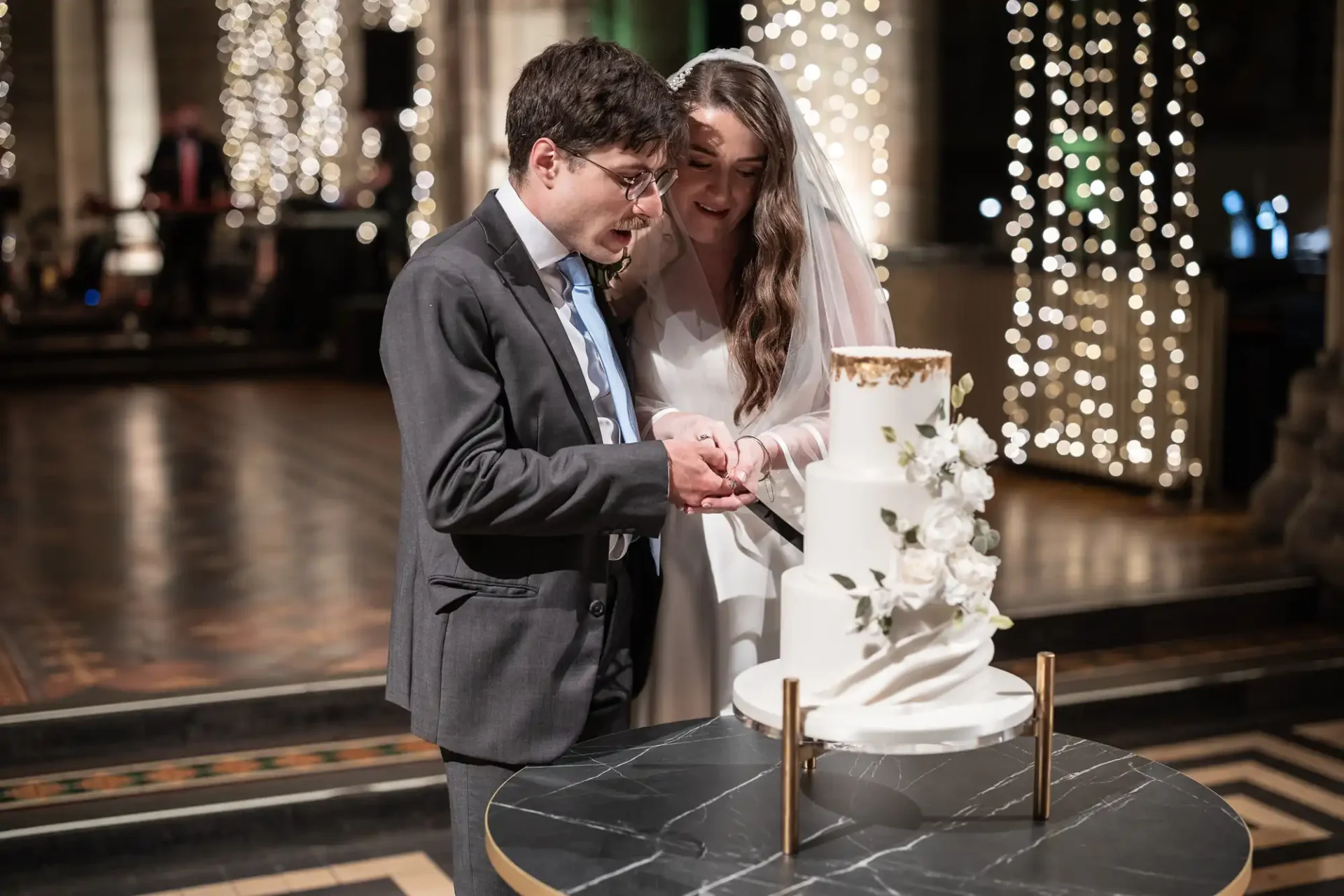 A bride and groom cut a tiered wedding cake adorned with white flowers, inside a venue decorated with string lights.