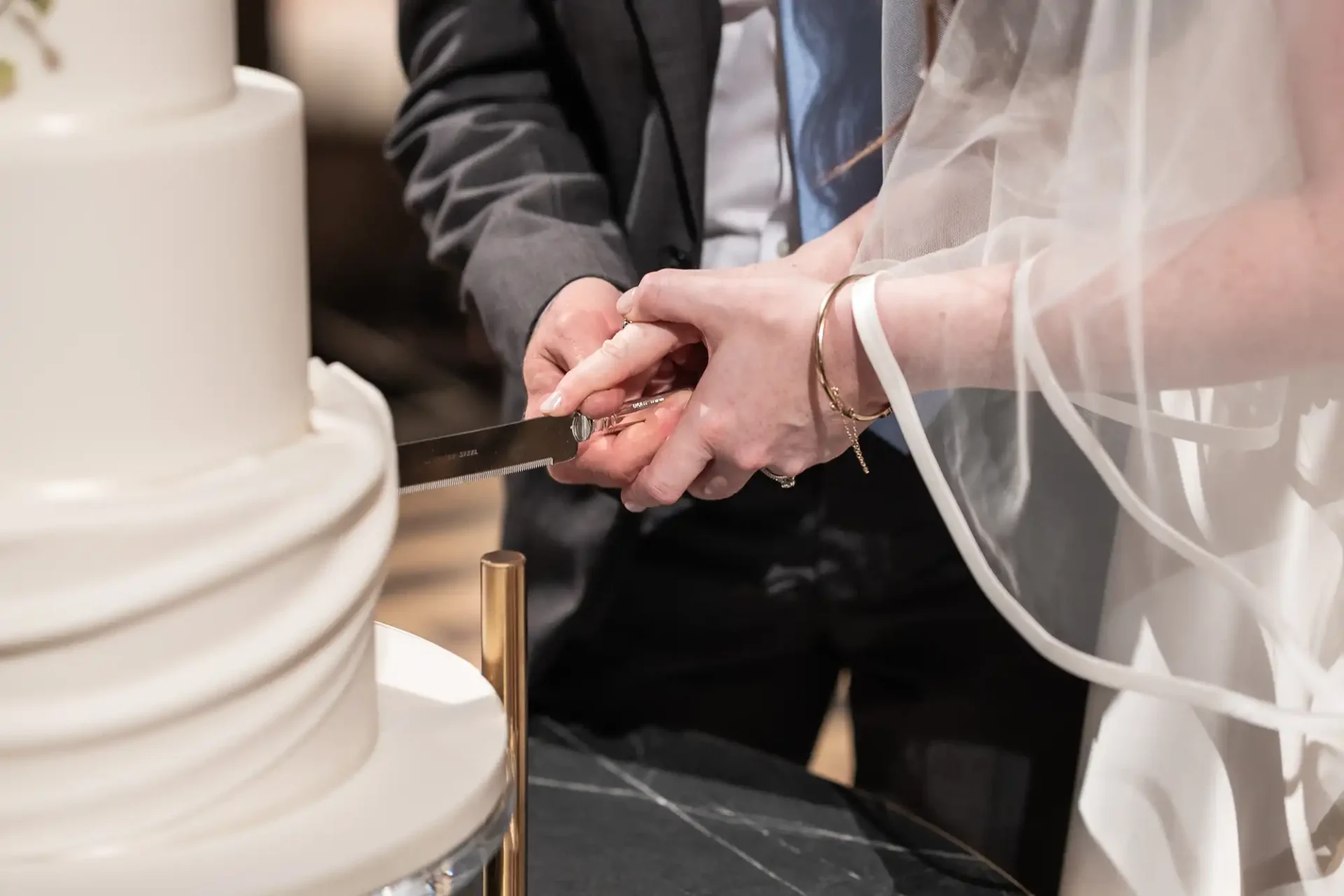 A couple cuts into a tiered white wedding cake together with a knife.