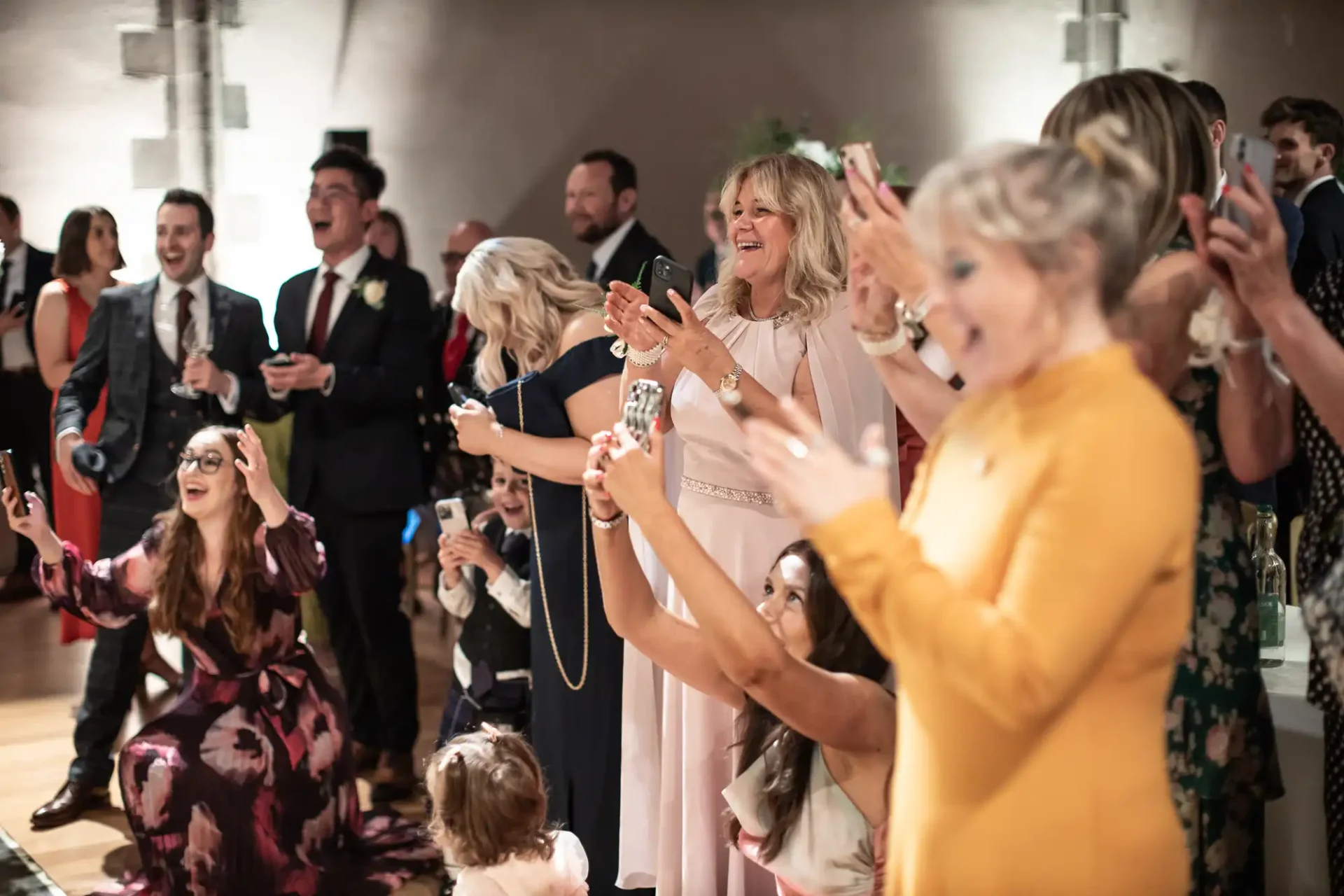 Group of people smiling and clapping at an indoor event. Women in dresses and men in suits are holding drinks and phones. A child is kneeling at the front.