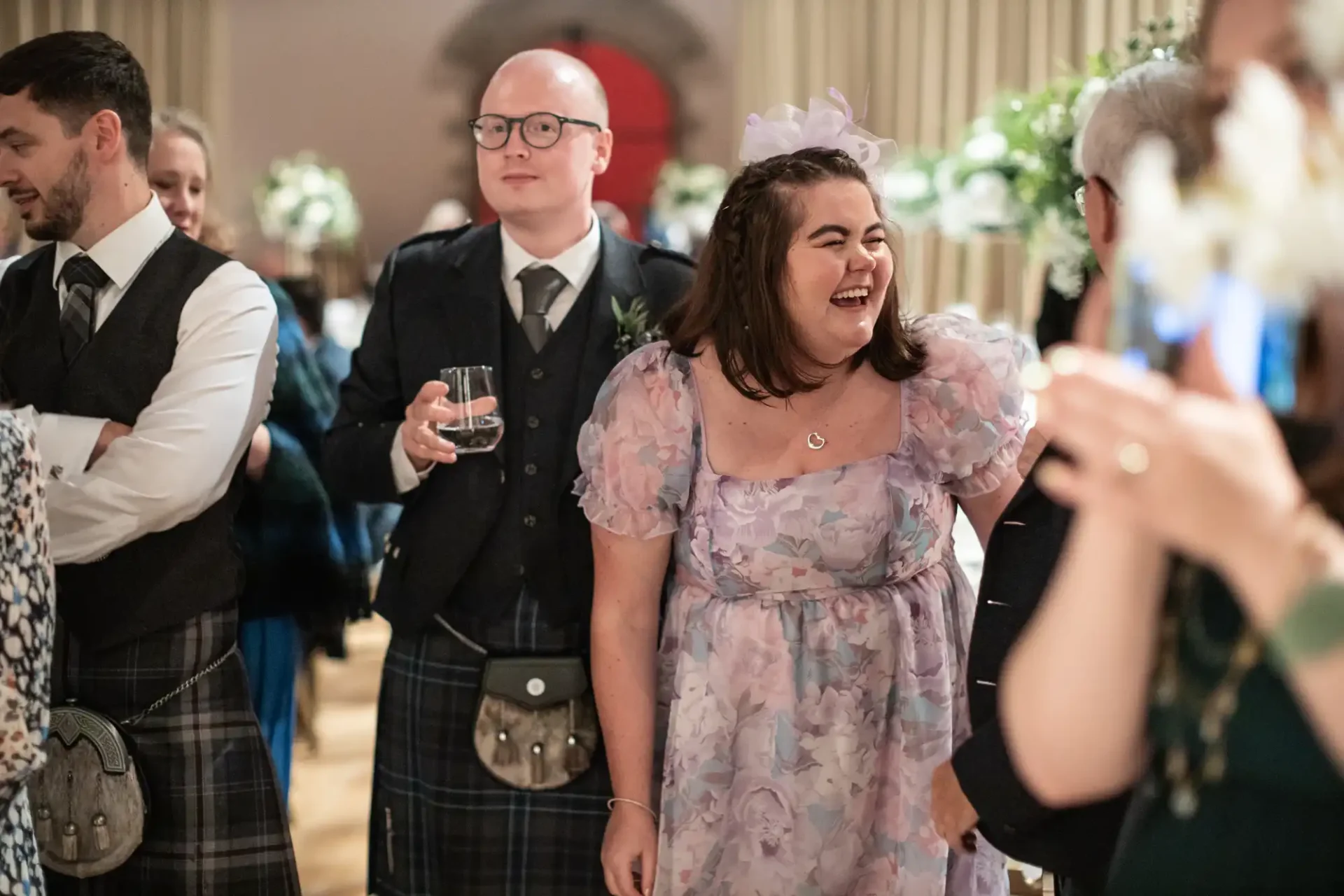 People dressed in formal attire, including kilts, at an indoor event. A woman in a floral dress laughs, while a man beside her holds a drink. Guests engage around them.