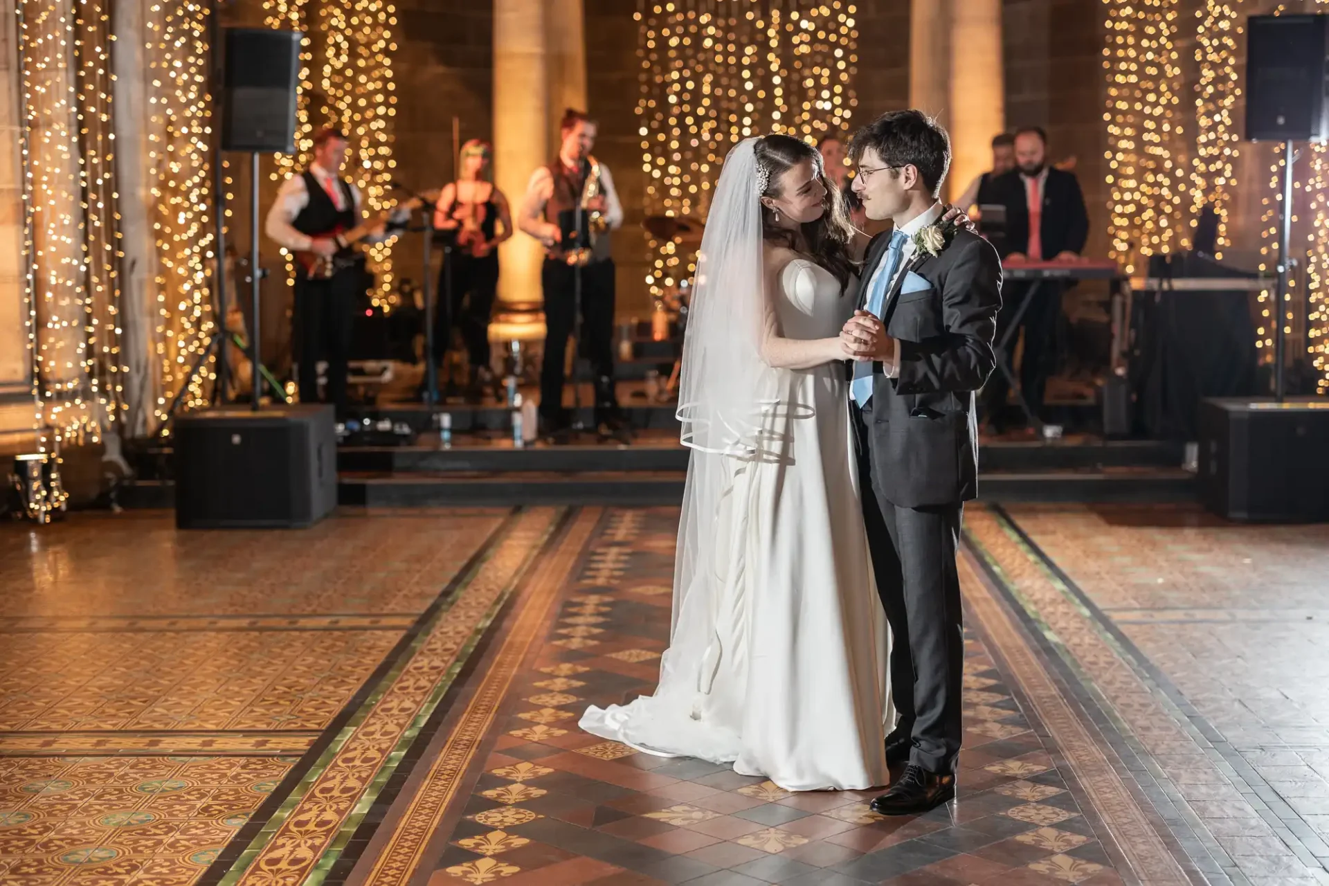 Bride and groom dance in an ornate hall, surrounded by fairy lights and musicians in the background.