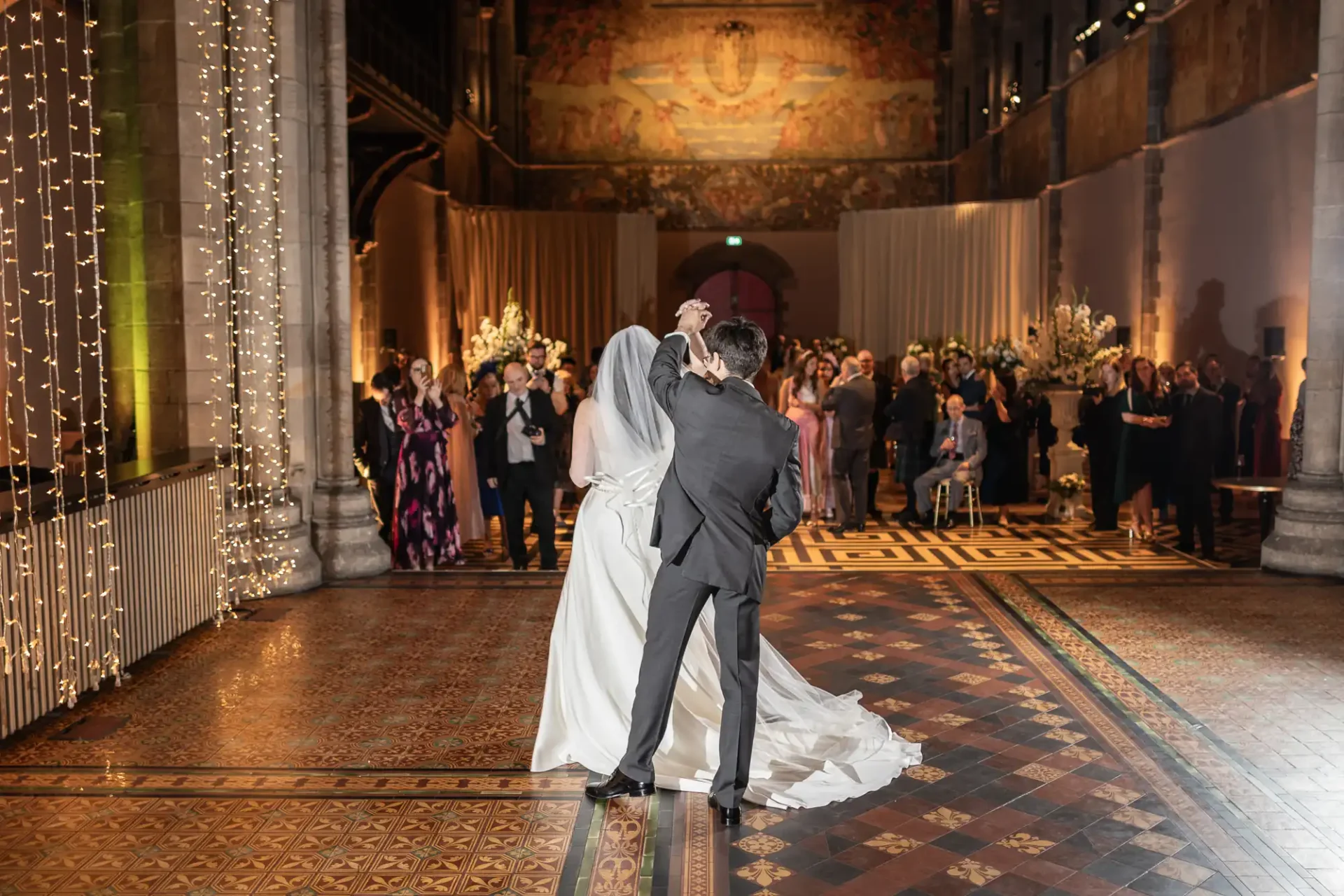 A bride and groom share their first dance on a patterned floor in a large, elegantly decorated hall filled with guests.