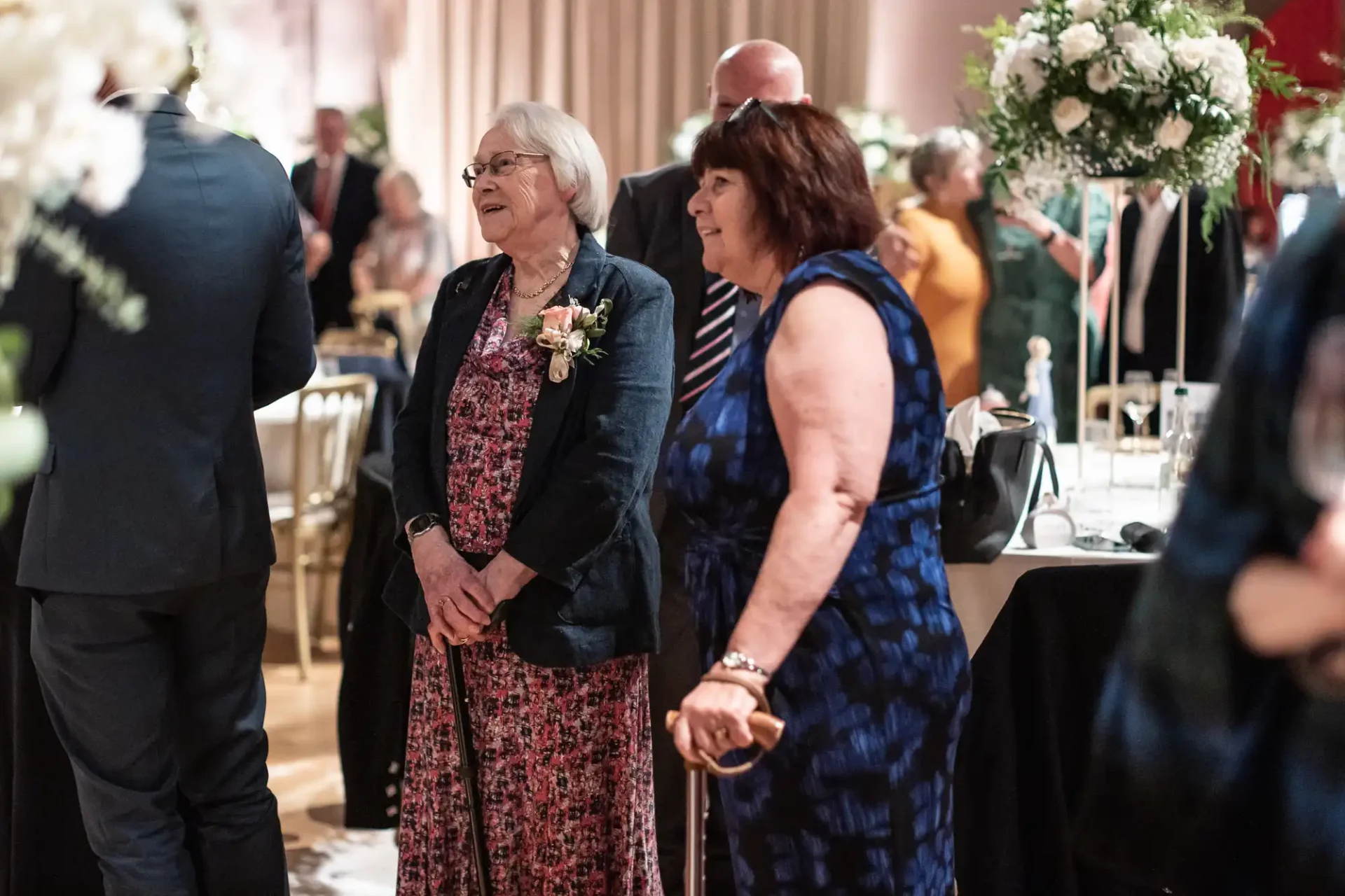 Two women stand and smile at an event, surrounded by elegantly set tables and floral decorations.