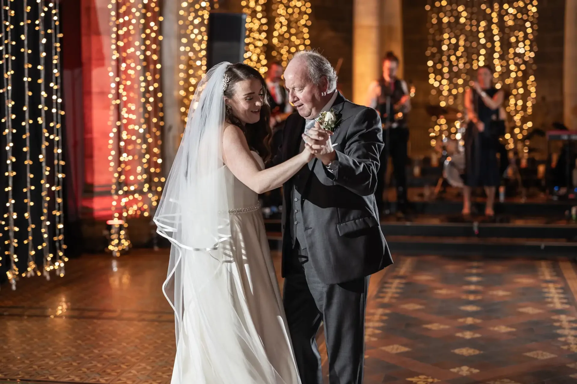 A bride and an older man, possibly her father, dance together at a wedding reception. They are surrounded by string lights and a band playing in the background.