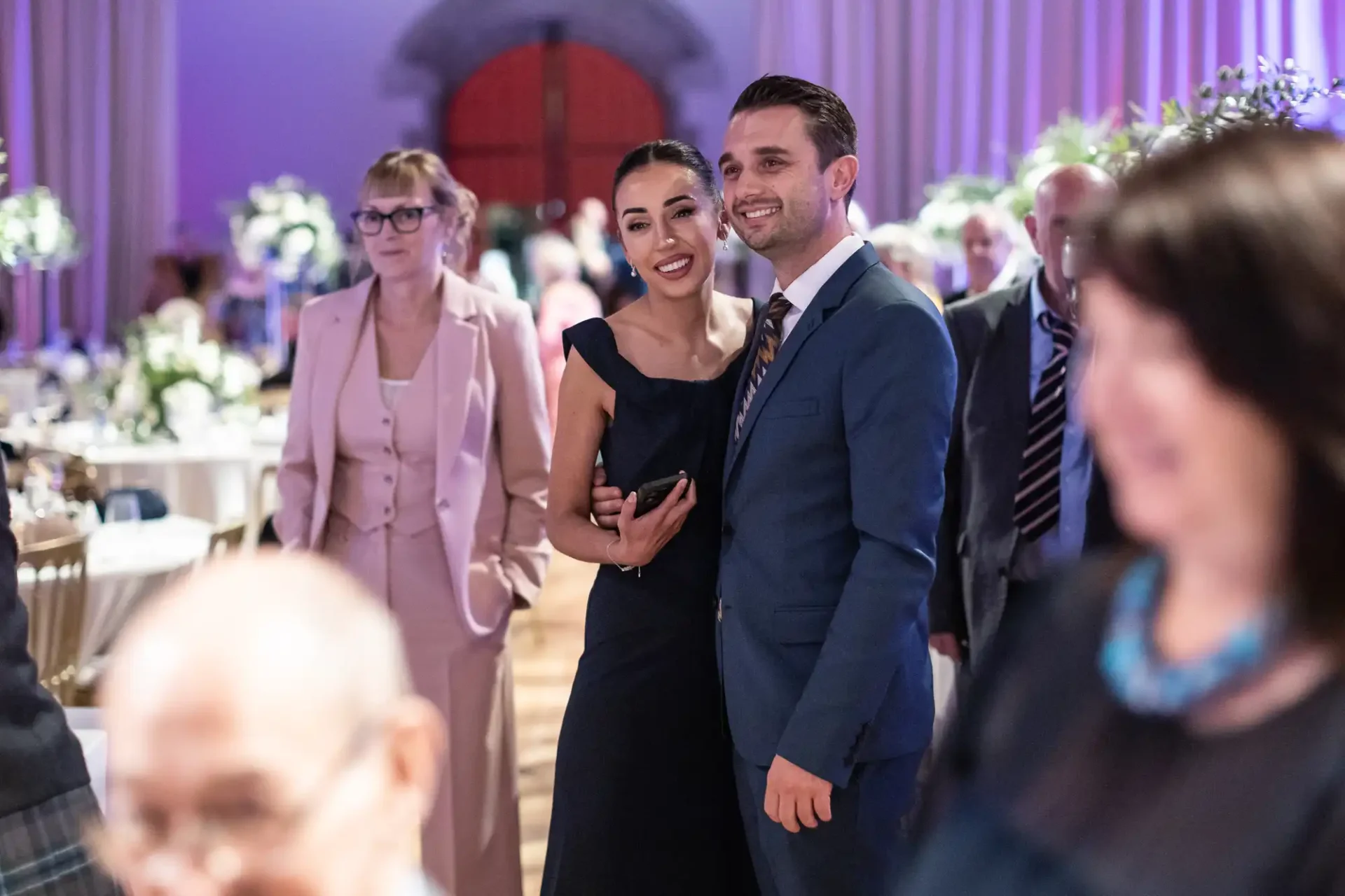 A couple in formal attire poses for a photo at a social gathering, surrounded by other attendees in an elegant setting with floral decorations.