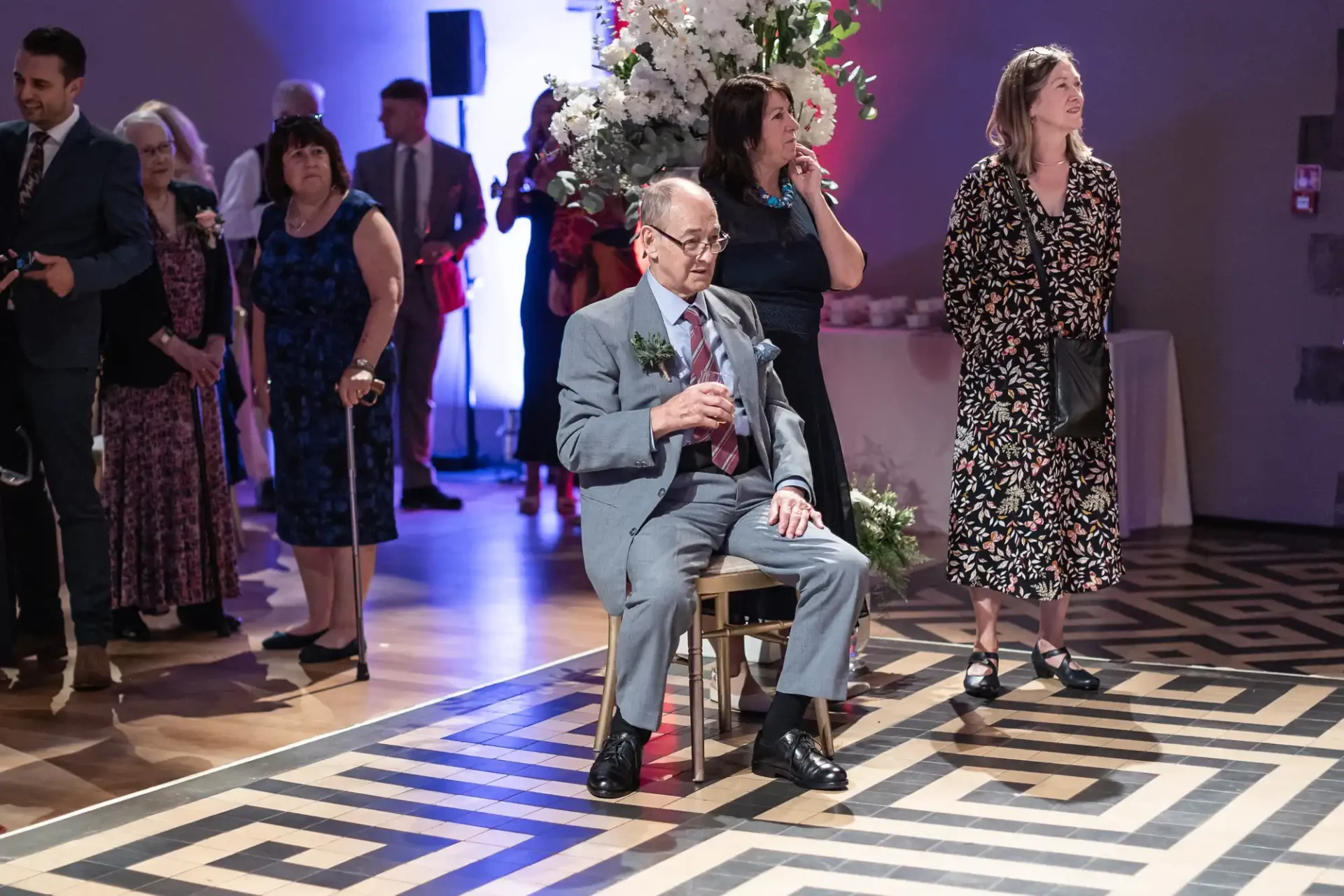 Elderly man in a suit sits on a chair holding a drink, surrounded by standing people at an indoor event with floral decor.