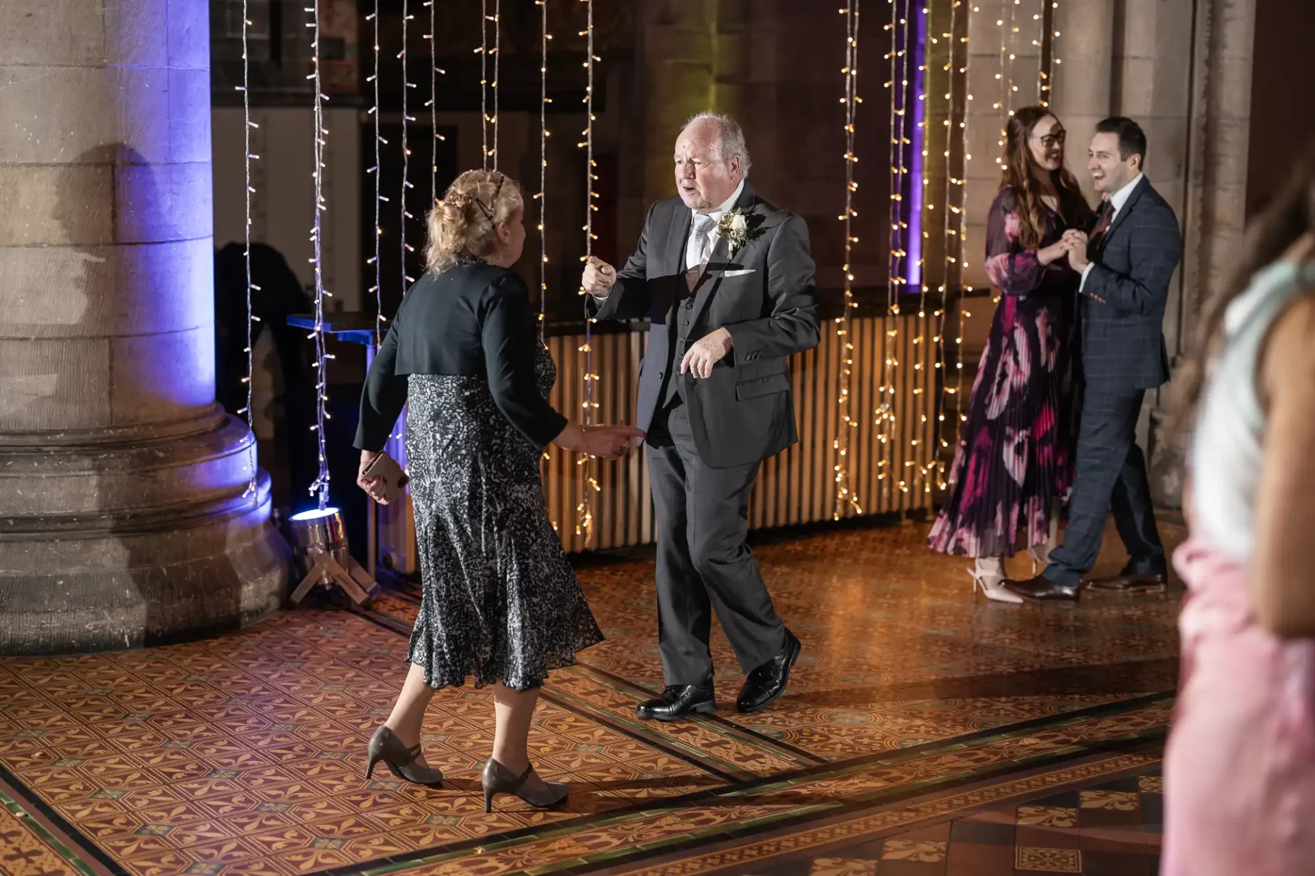 A man and a woman dance on a patterned floor in front of string lights. Two people smile and watch in the background.