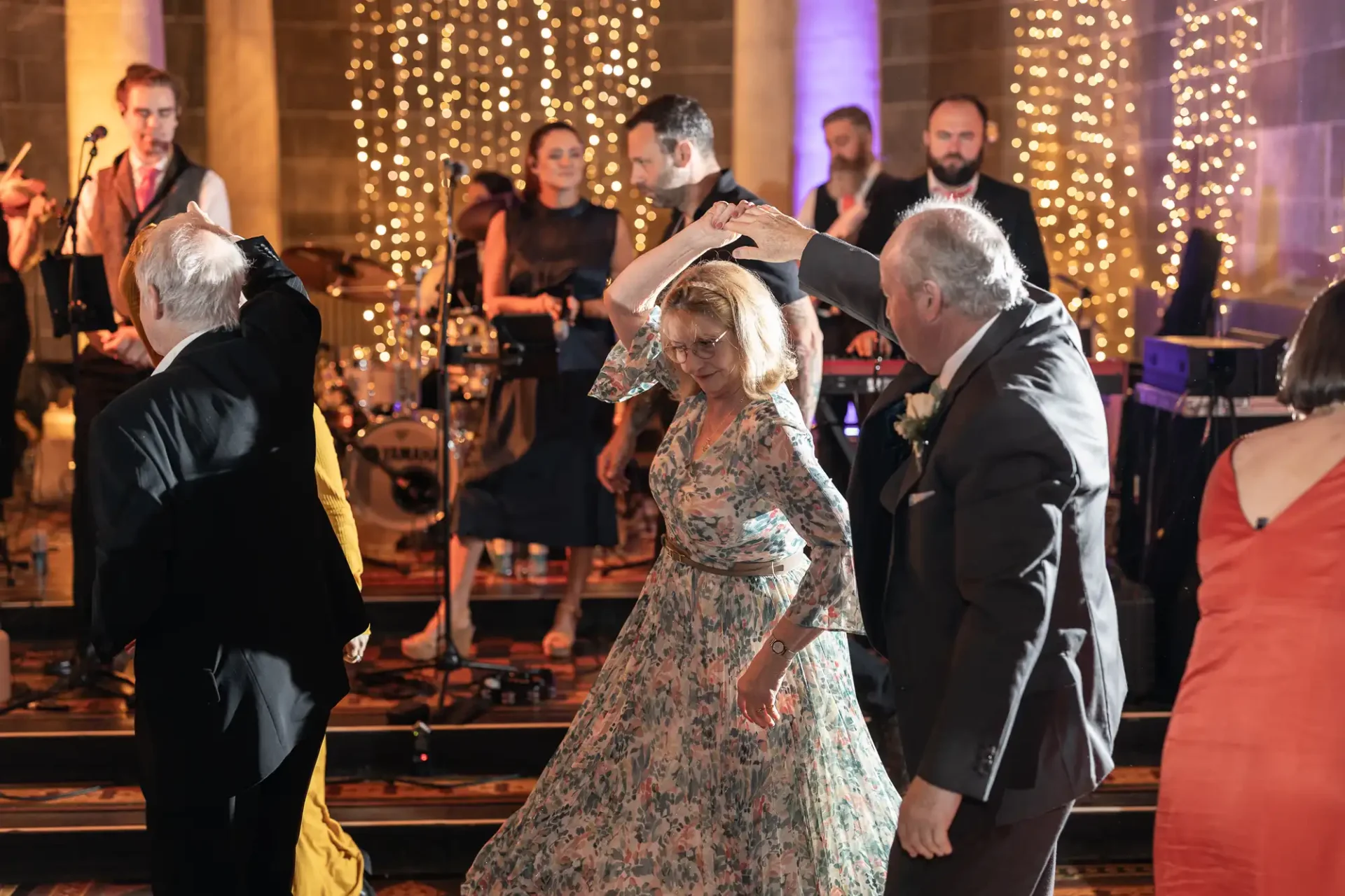 A couple in formal attire dances at an event, with a live band and audience in the background. String lights and elegant decor set the atmosphere.