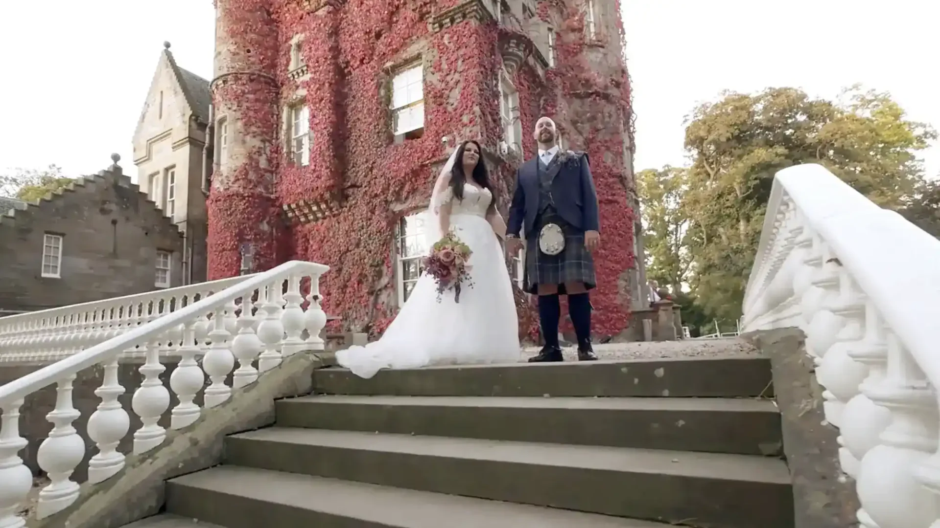 wedding video at Carlowrie Castle: A bride in a white gown and a groom in a kilt stand on stone steps in front of a large, ivy-covered building with ornate architecture.