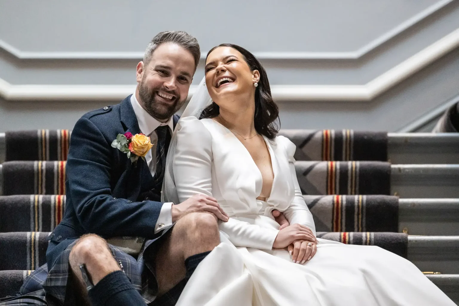A bride and groom sit on carpeted stairs, smiling and laughing together after being married in Edinburgh. The groom wears a dark suit with a floral boutonniere; the bride wears a white dress with a deep neckline.