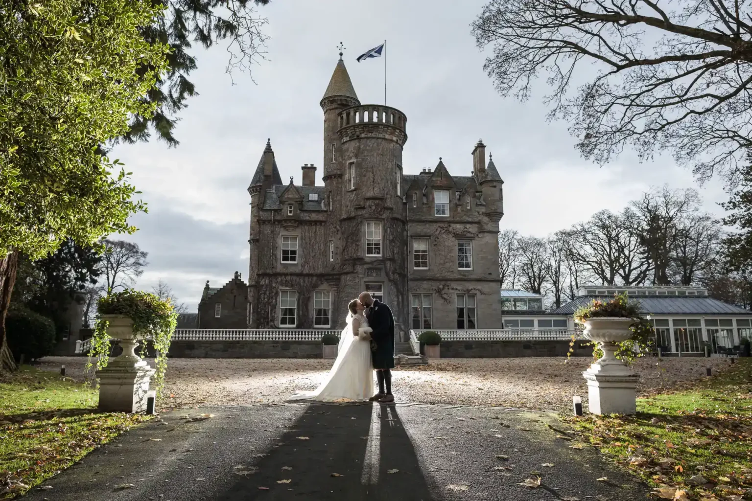 Wedding photographer in Edinburgh using backlight to photograph a bride and groom standing together in front of a Carlowrie Castle with a Scottish flag, surrounded by trees and formal gardens.