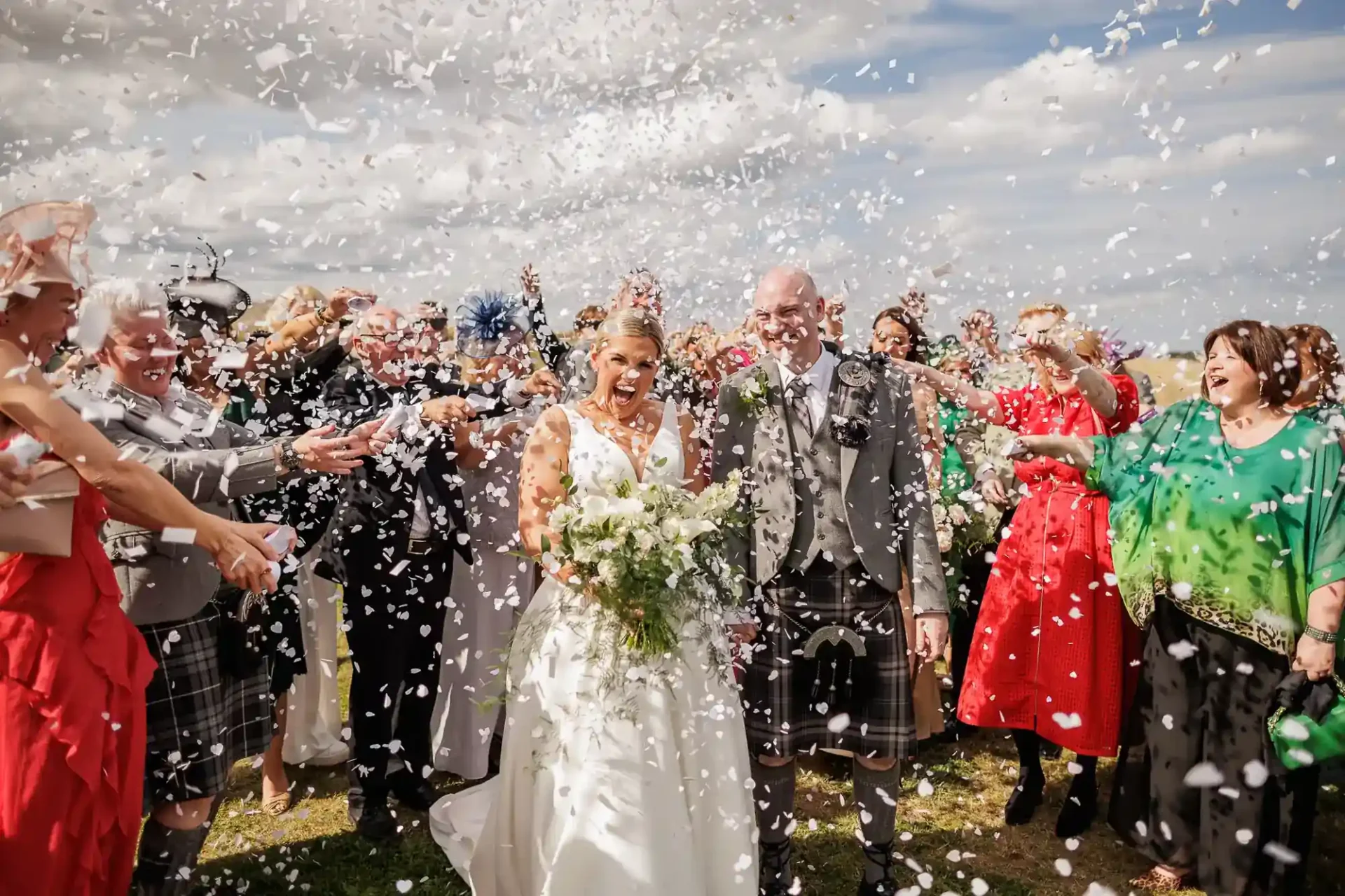 A bride in a white dress and a groom in a grey jacket and kilt smile as guests shower them with white confetti outdoors during their wedding at Whitekirk Hill. The crowd is joyful, dressed in colourful outfits, under a cloudy sky. The mood is festive and celebratory.