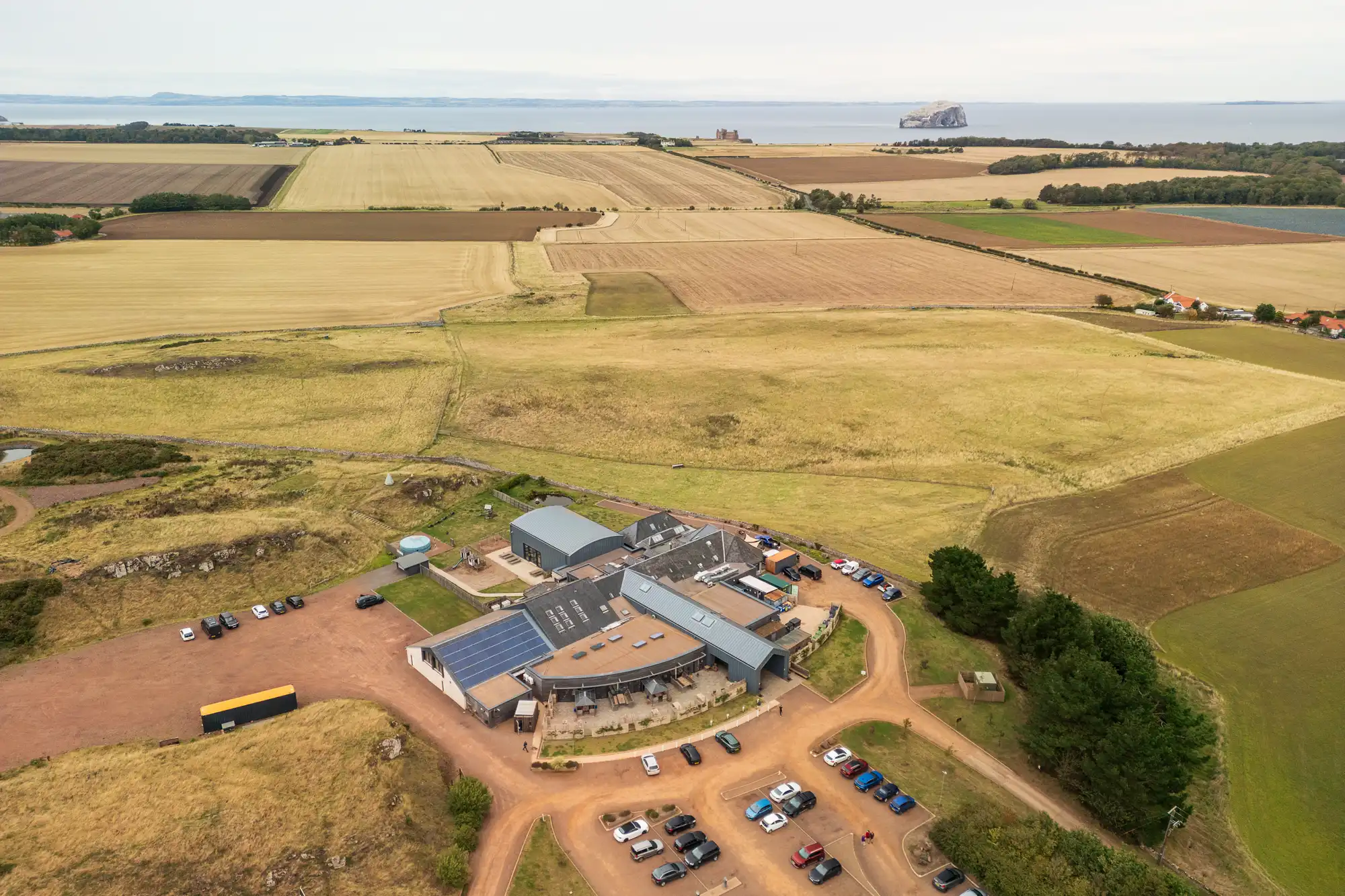 Aerial view of a countryside building complex with multiple roofs, outdoor seating, and car park surrounded by fields. Several cars are parked; patchwork farmland stretches to the horizon. The sea and a rocky island are visible in the background under a cloudy sky.
