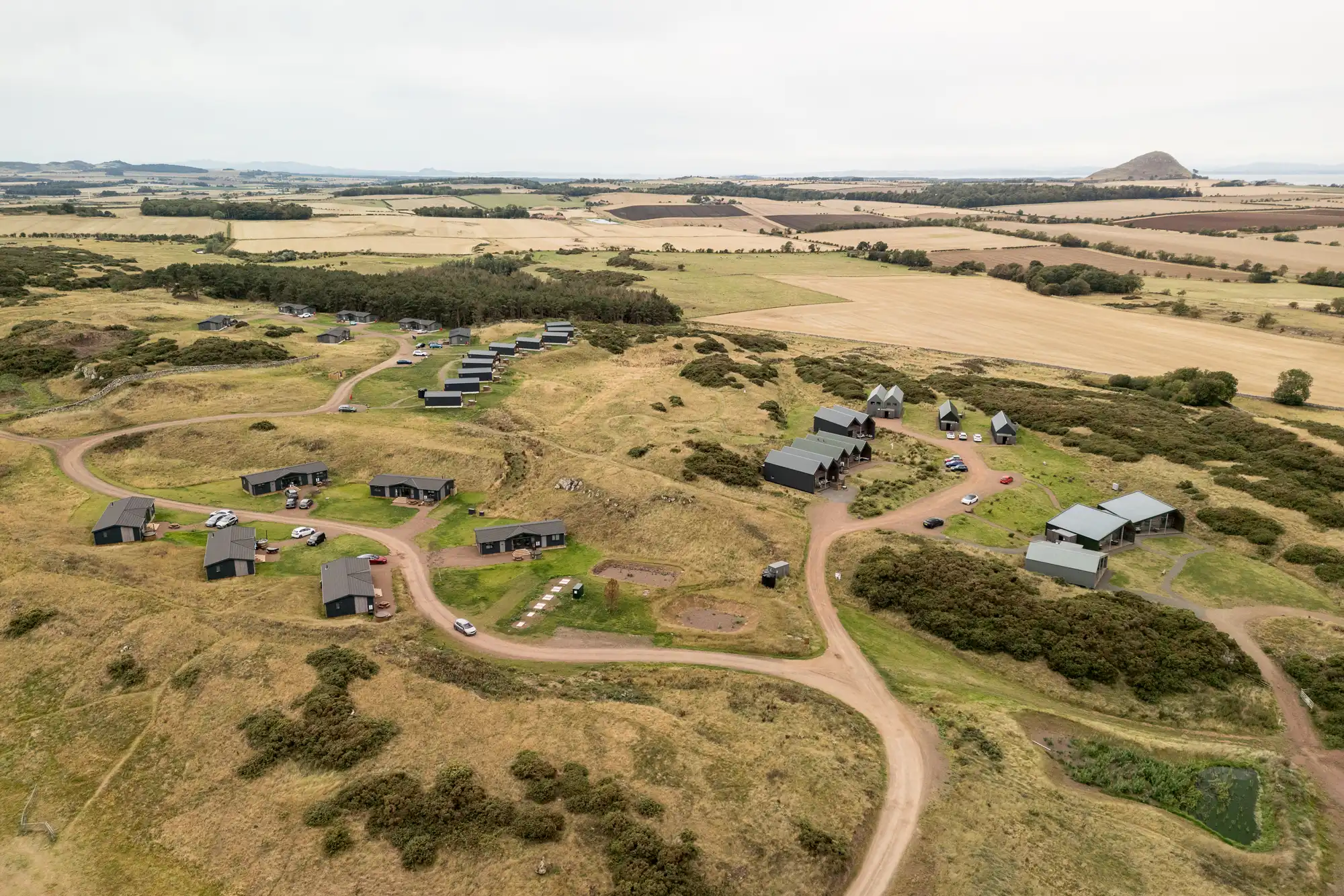 Aerial view of a rural landscape with scattered modern cabins, winding dirt roads, grassy hills, and farmland. Clustered buildings are surrounded by fields, trees, and small ponds, with rolling countryside stretching into the distance under an overcast sky.