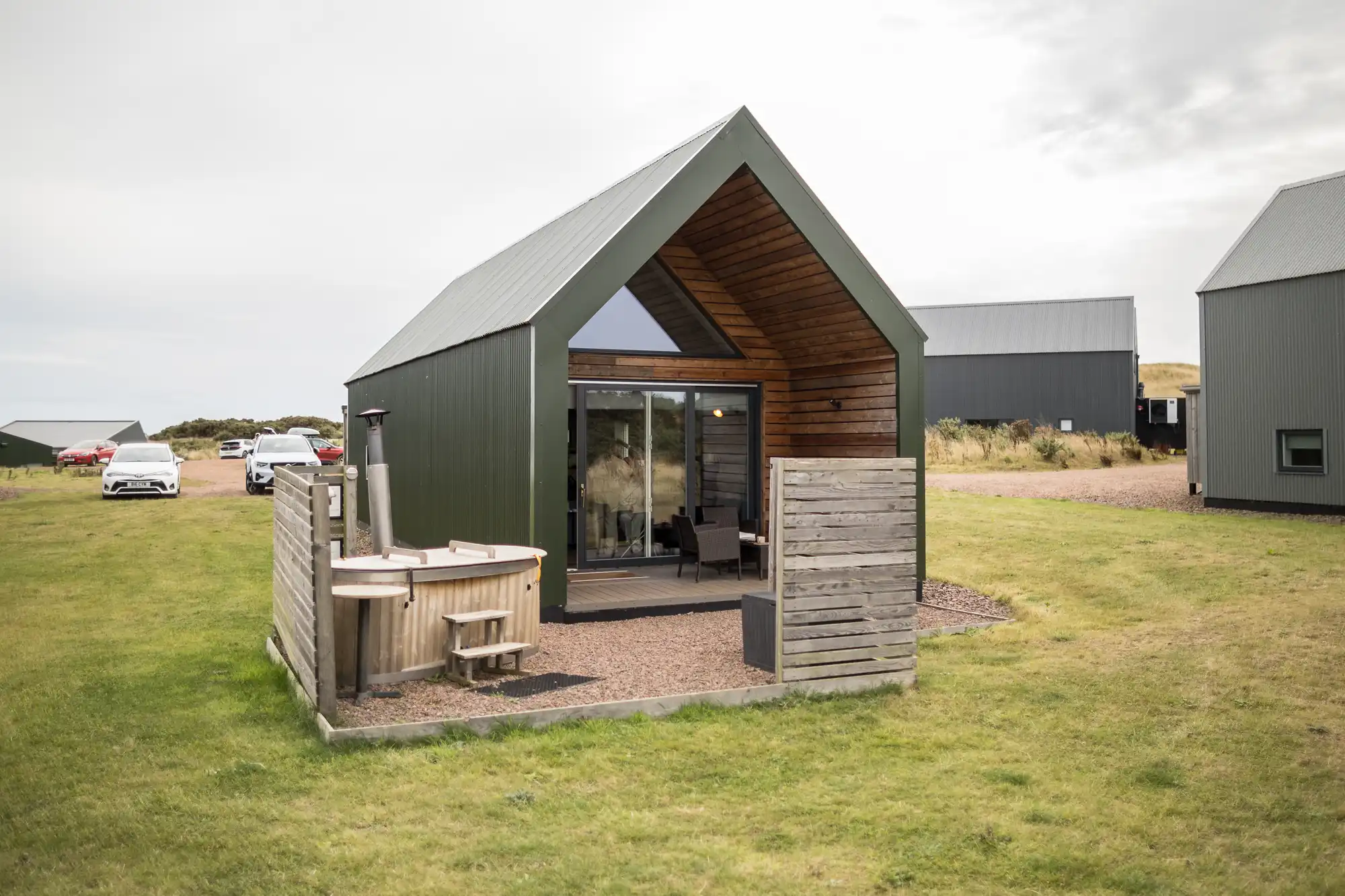 A modern, dark green cabin with an open wooden veranda and glass doors sits on a grassy field. There’s a small outdoor fenced area with a wooden hot tub. Other similar cabins and parked cars are visible in the background under a cloudy sky.