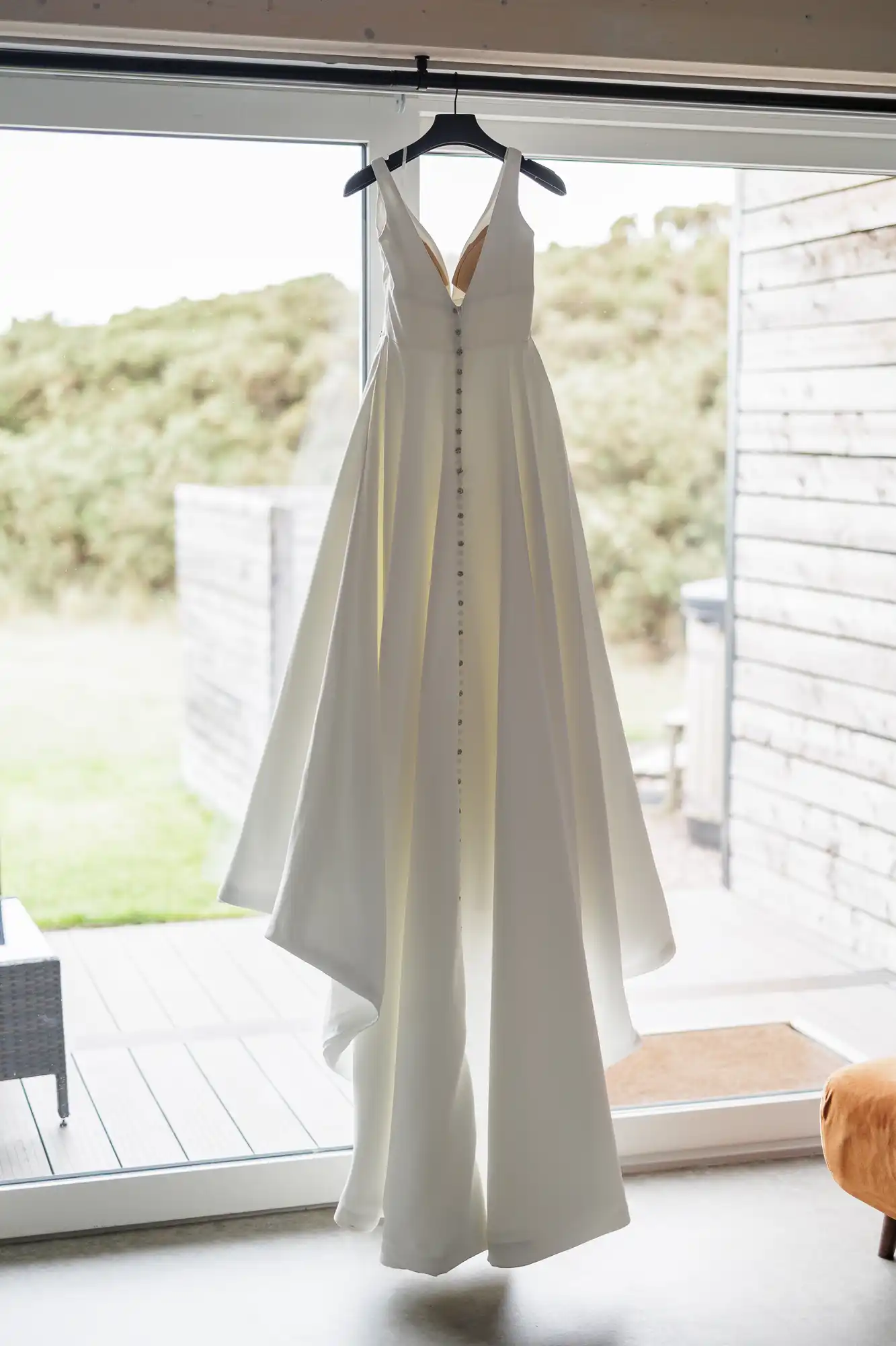 A white, sleeveless wedding dress with a plunging neckline, fitted bodice, and full skirt hangs on a black hanger indoors. The dress features a row of buttons down the front and is displayed near a large window, with greenery visible outside.