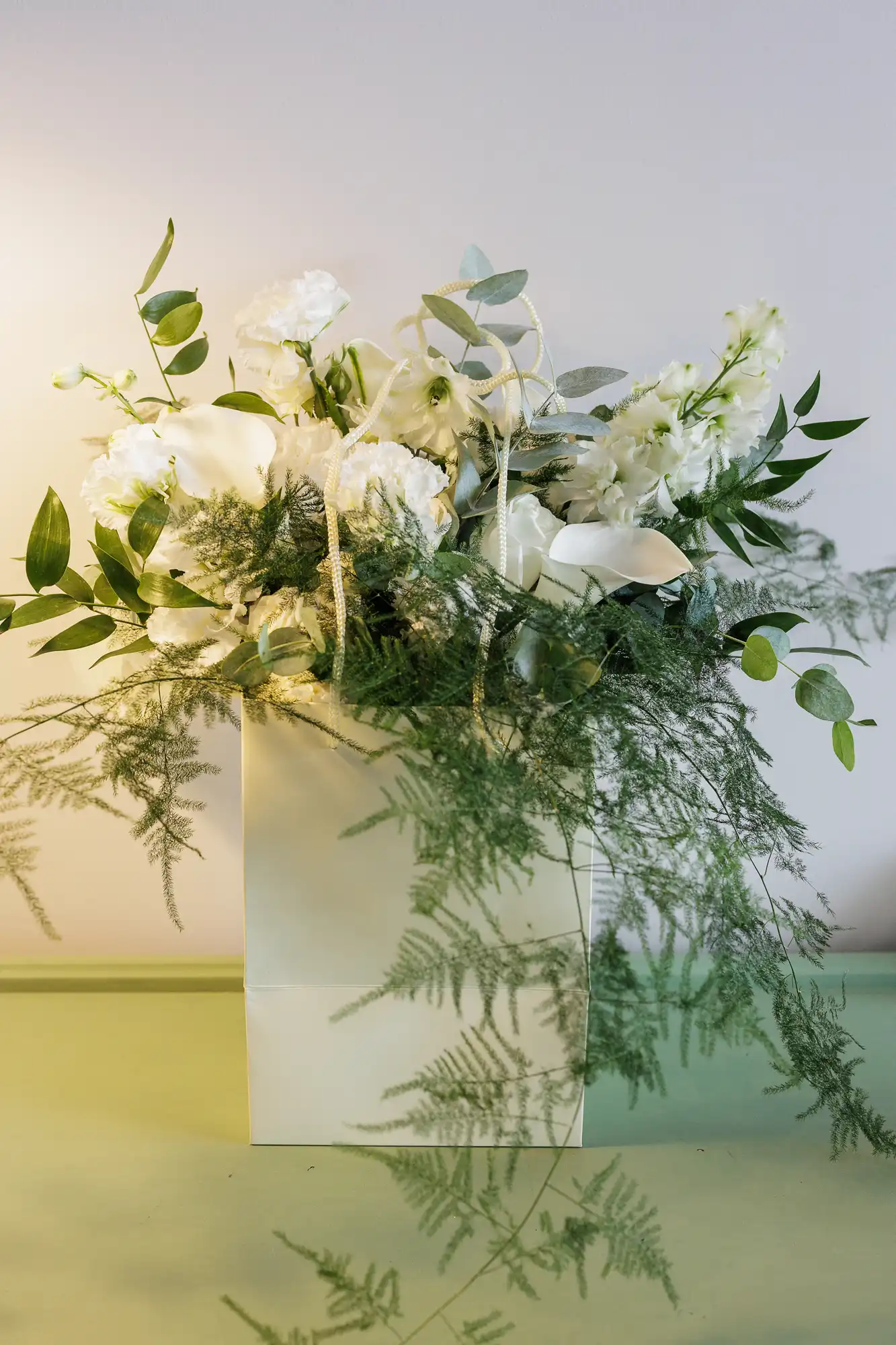 A white paper bag holds an elegant bouquet of white flowers, green leafy branches, and fern fronds, arranged against a plain pale background. The foliage and flowers spill gracefully over the edges of the bag.