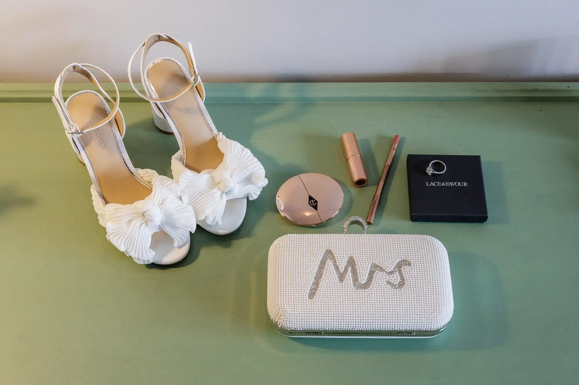 A pair of white high-heeled sandals with bows, a pearl-studded clutch with "Mrs" embroidered, rose gold lipstick, powder compact, eyeliner pencil, and a black box with a ring, all arranged neatly on a pale green surface.