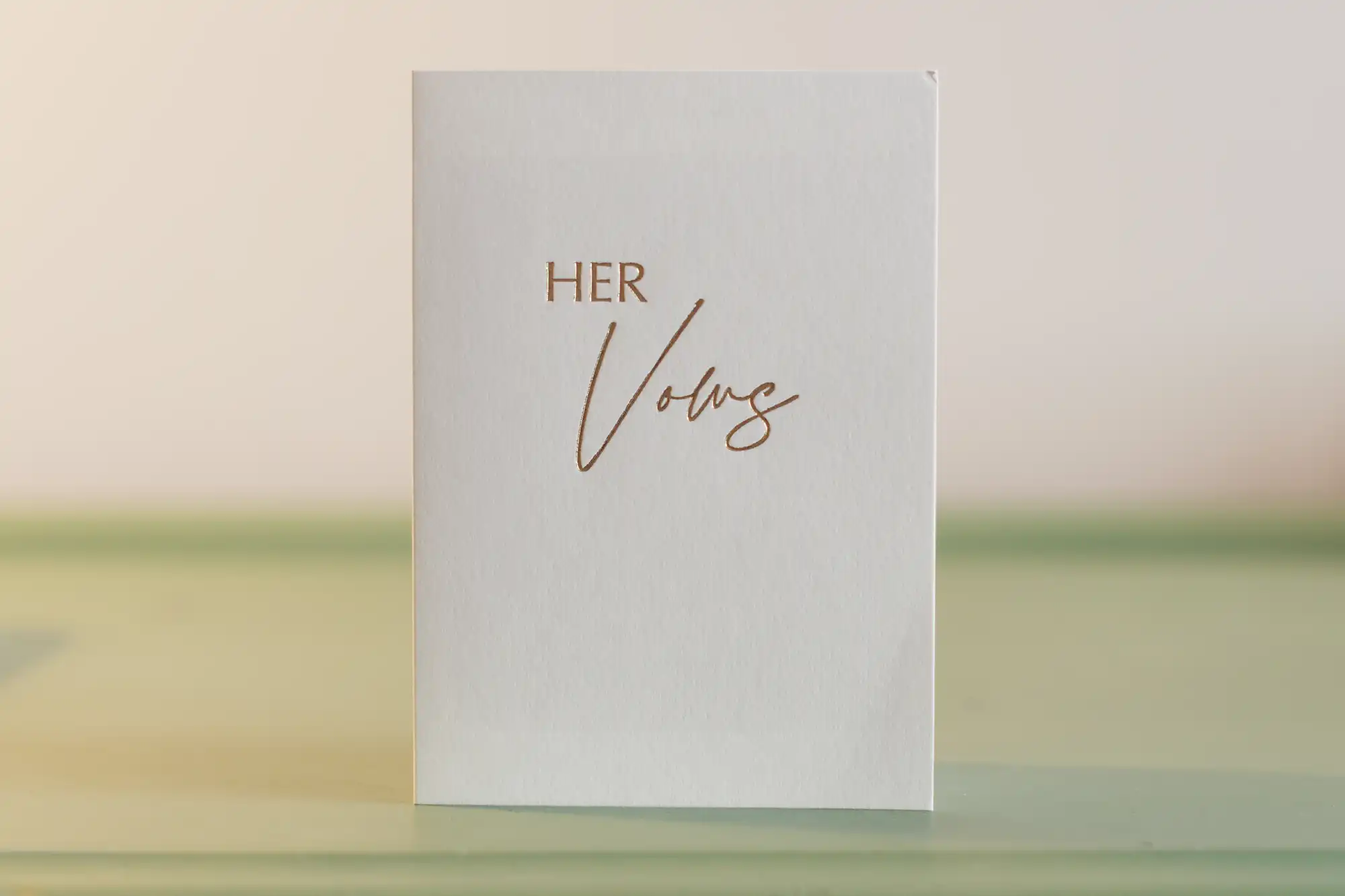 A white card stands upright on a pale green surface. The words "HER Vows" are written on the front in elegant gold script and uppercase lettering. The background is simple and softly blurred.