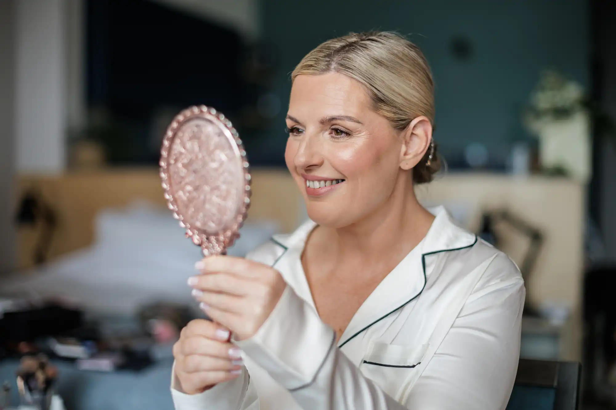 A smiling woman with blonde hair, wearing silky white pyjamas with dark piping, holds an ornate hand mirror, looking at her reflection. The background is softly blurred, showing a bed and some room decor. The atmosphere appears calm and relaxed.
