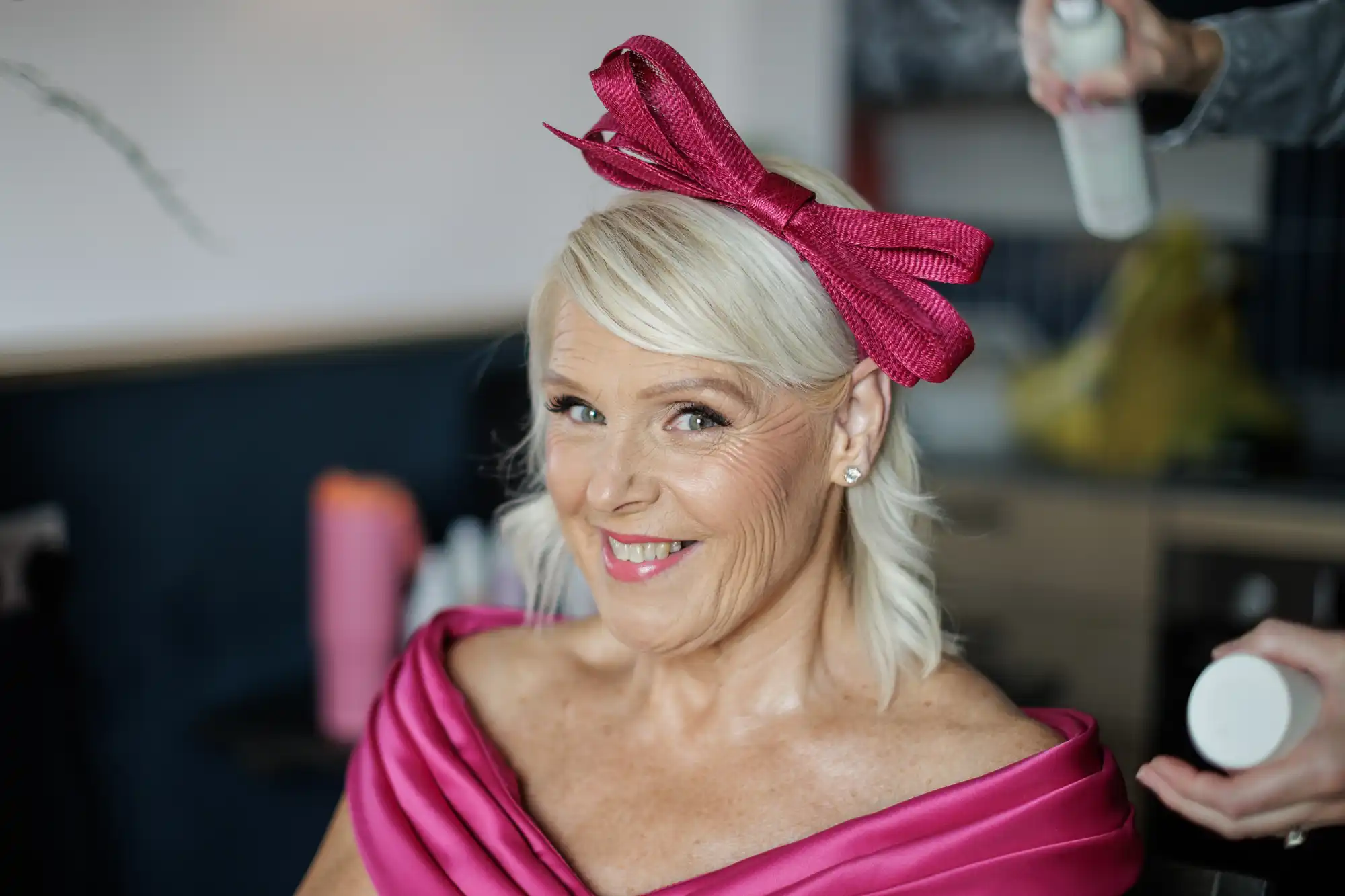 A smiling older woman with short blonde hair wears a bright pink dress and a large matching pink bow on her head. She is seated indoors, looking at the camera, while someone sprays her hair with hairspray. The background is softly blurred.