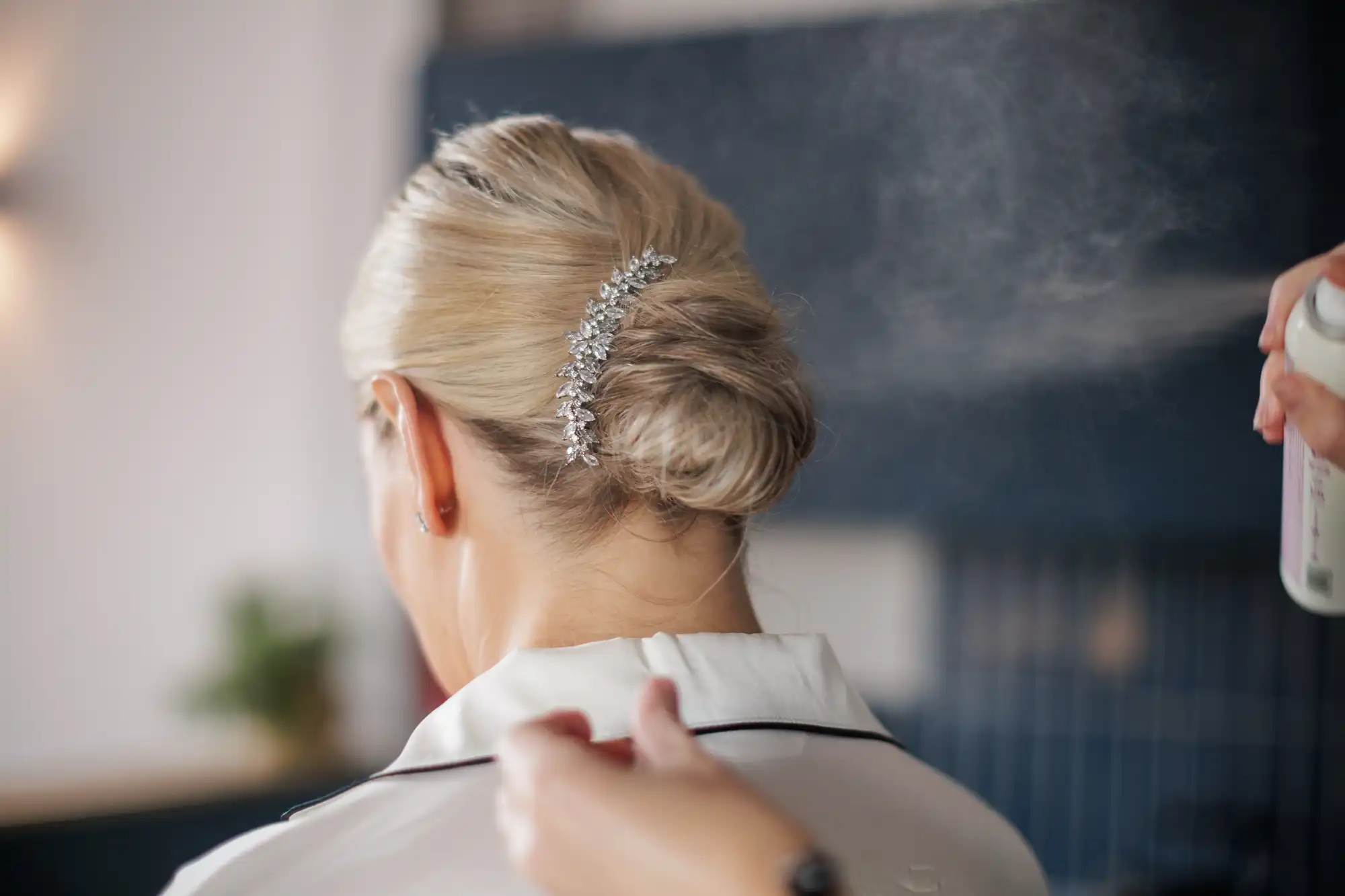 A woman with blonde hair styled in an elegant low bun adorned with a decorative silver hairpiece is seen from behind. Someone is spraying hairspray onto her hair, and she is wearing a light-coloured top with a dark collar. The background is softly blurred.