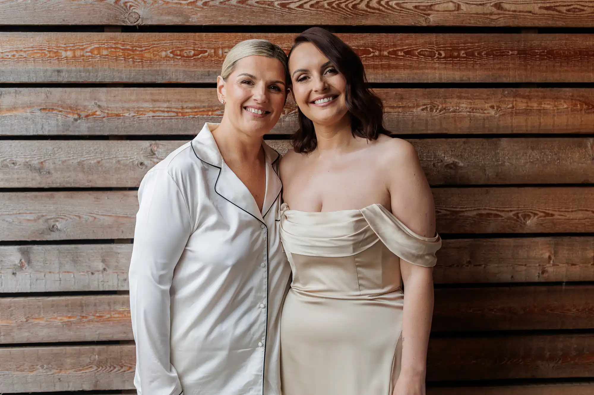 Two women stand close together smiling in front of a wooden plank wall. One wears a silky white pyjama-style top; the other wears an off-the-shoulder, cream-coloured dress with draping. Both have light skin and short hair, one blonde and one brunette.