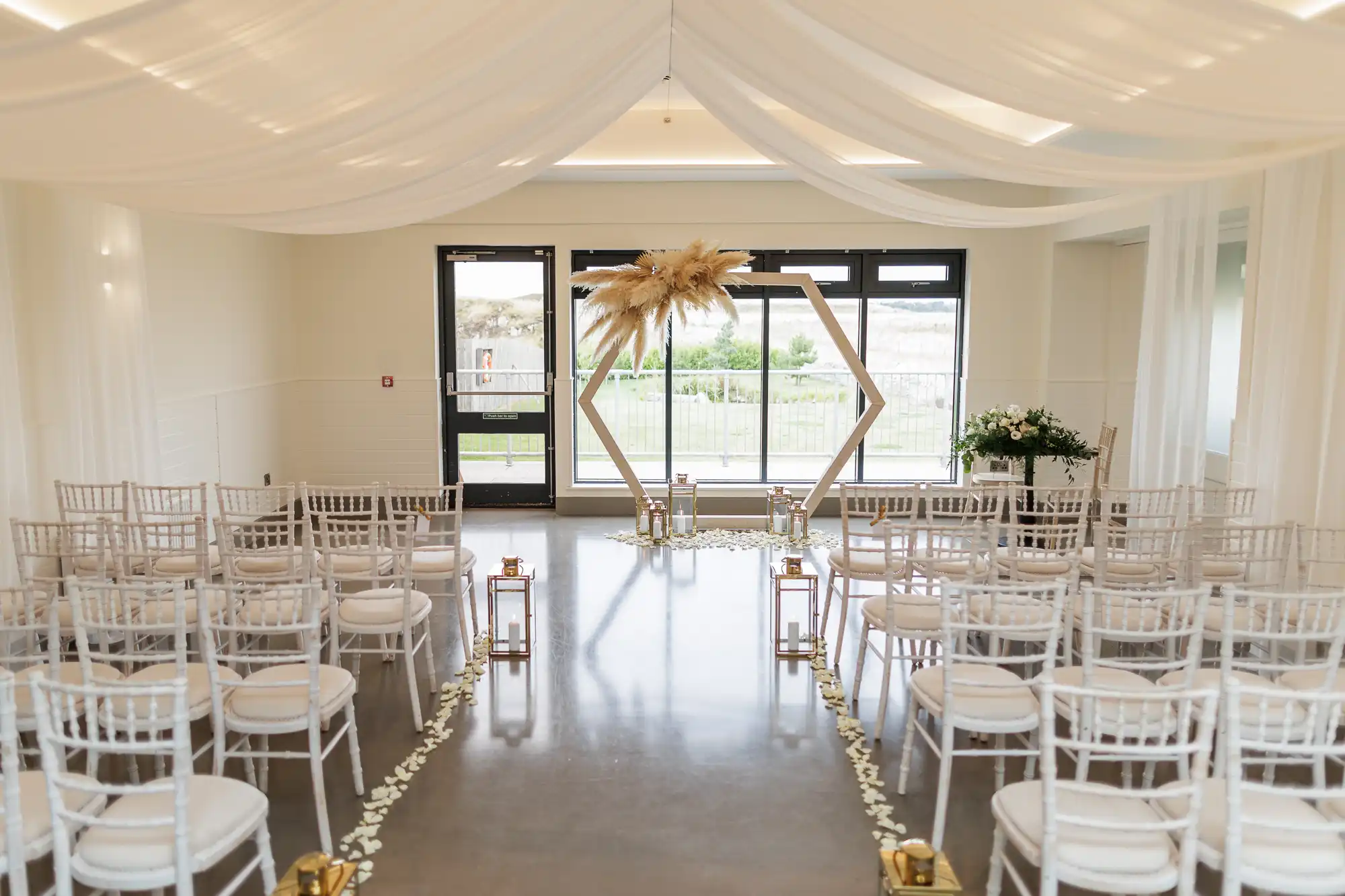 A bright, modern wedding ceremony setup with white chairs arranged in rows facing a hexagonal arch decorated with pampas grass. White drapes hang from the ceiling, petals line the aisle, and gold lanterns are placed along the aisle edges. Large windows are at the back.