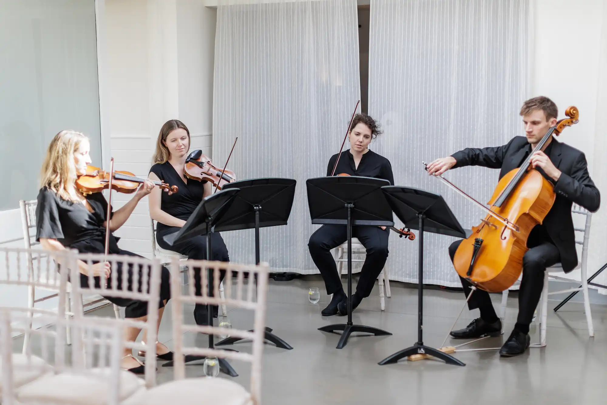 Four musicians dressed in black perform indoors as a string quartet. Three play violins or violas, and one plays a cello. They are seated in a semi-circle with music stands in front of them. The bright room has pale chairs and sheer curtains in the background.