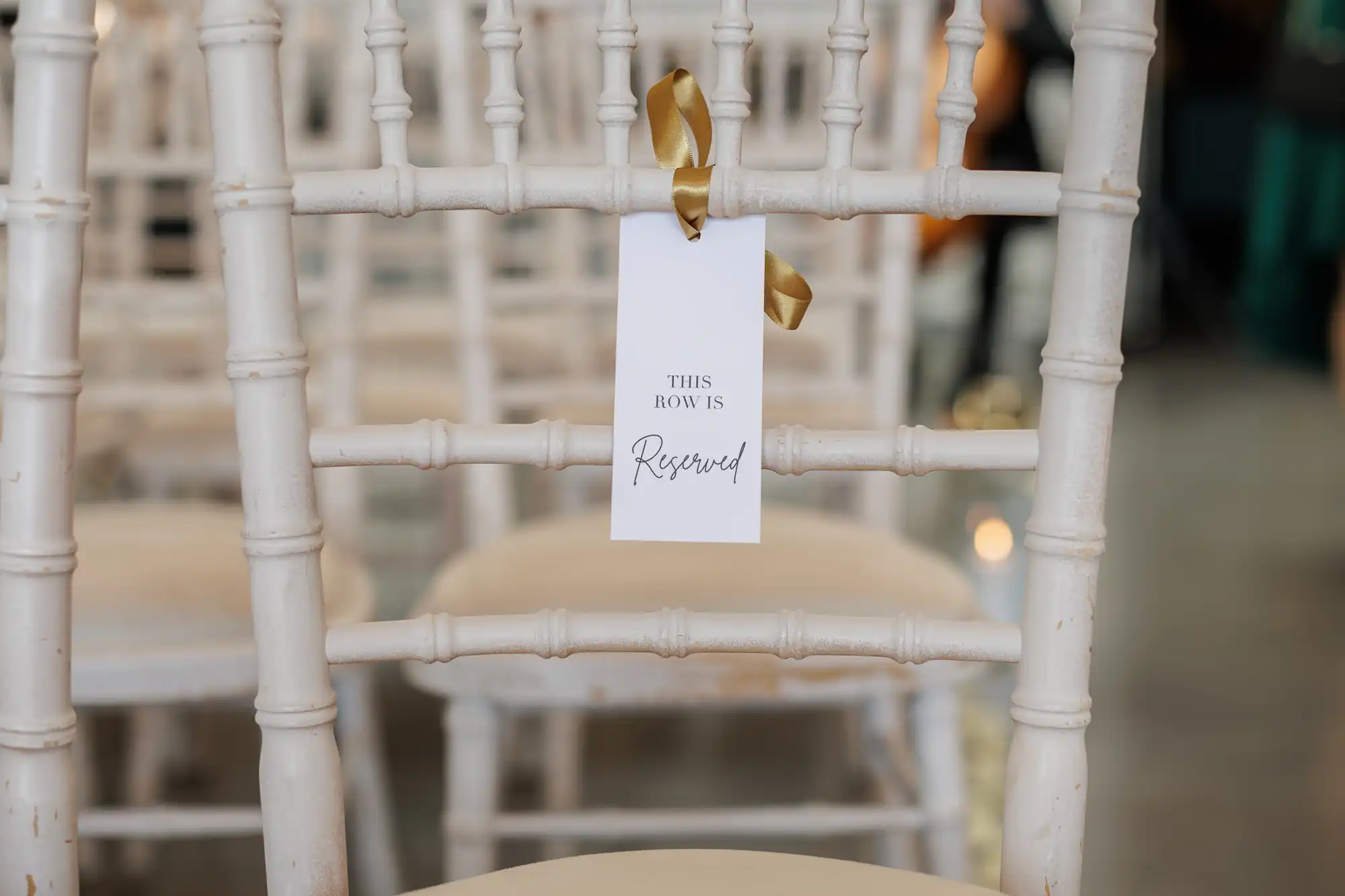 A white wooden chair with a beige cushion has a white label tied with a gold ribbon to its backrest. The label reads "THIS ROW IS Reserved" in elegant script, indicating the seat is reserved, with similar chairs in the background.