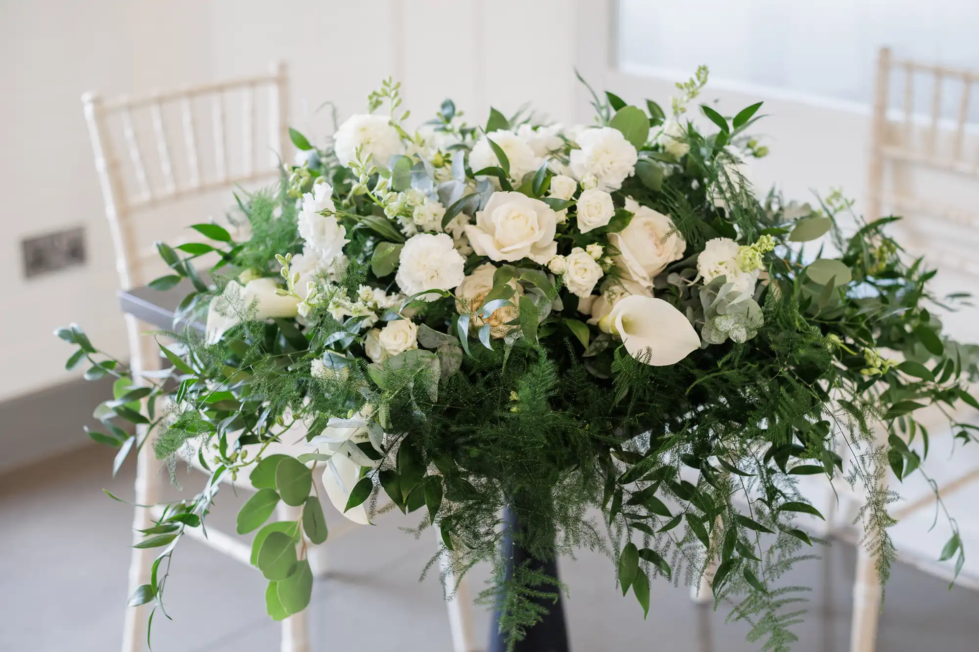 A floral arrangement of white roses, calla lilies, and assorted greenery decorates a table with white chairs behind it. The flowers and leaves cascade elegantly over the table edge in a bright, minimal indoor setting.