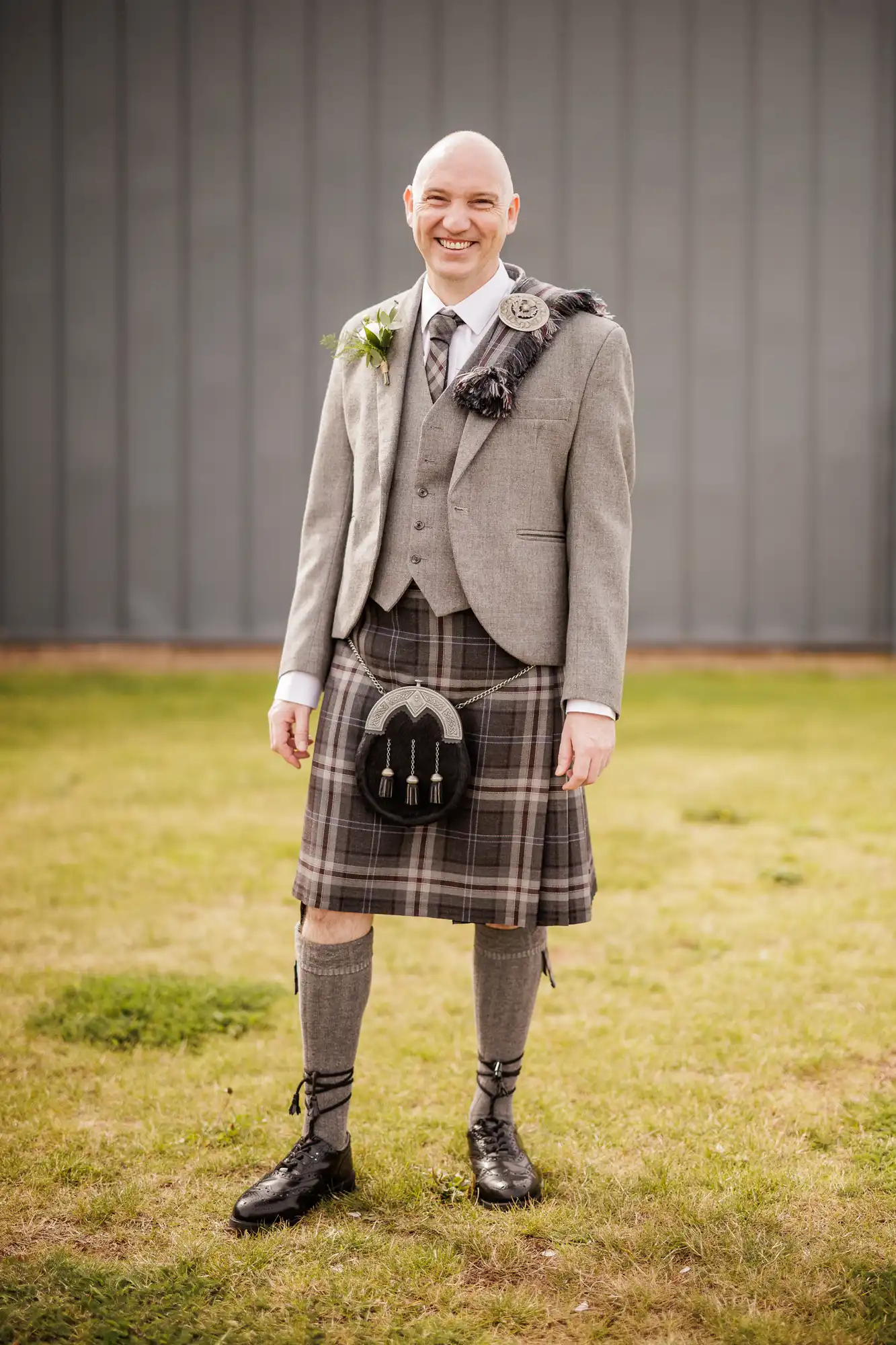 A smiling man stands on grass, wearing traditional Scottish attire including a grey jacket, waistcoat, tartan kilt, sporran, knee-high socks with flashes, and black lace-up shoes. He has a buttonhole and tartan sash over one shoulder. A grey wall is in the background.