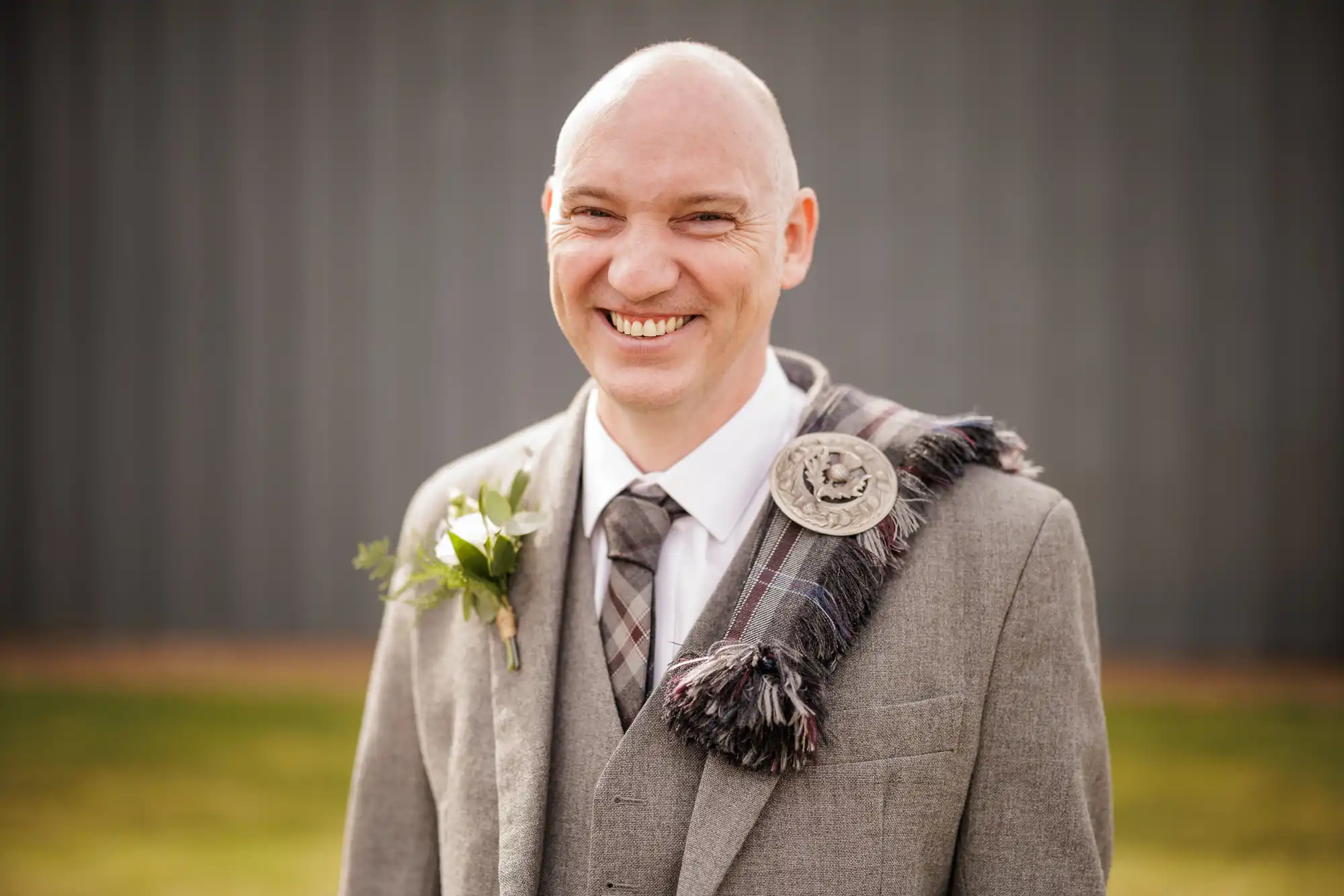 A smiling man with a bald head wears a grey suit, checked tie, and tartan sash with a silver brooch. He has a buttonhole pinned to his lapel. The background is blurred, showcasing a grassy area and a grey wall.