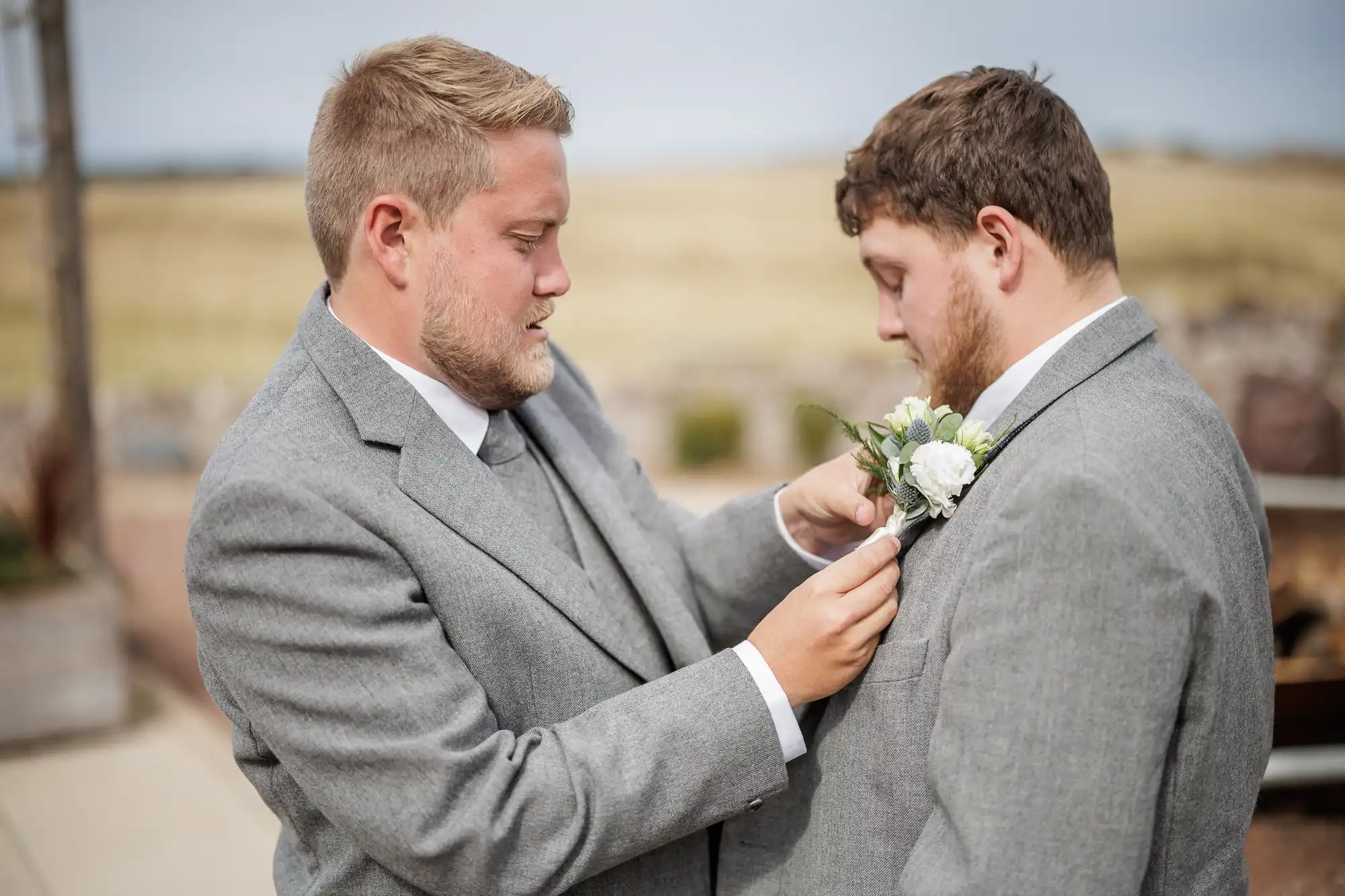 Two men in grey suits stand outside. One man pins a white flower buttonhole to the other’s lapel. Both have short hair and beards. The background is blurred, showing fields and a cloudy sky, suggesting a rural or outdoor setting.