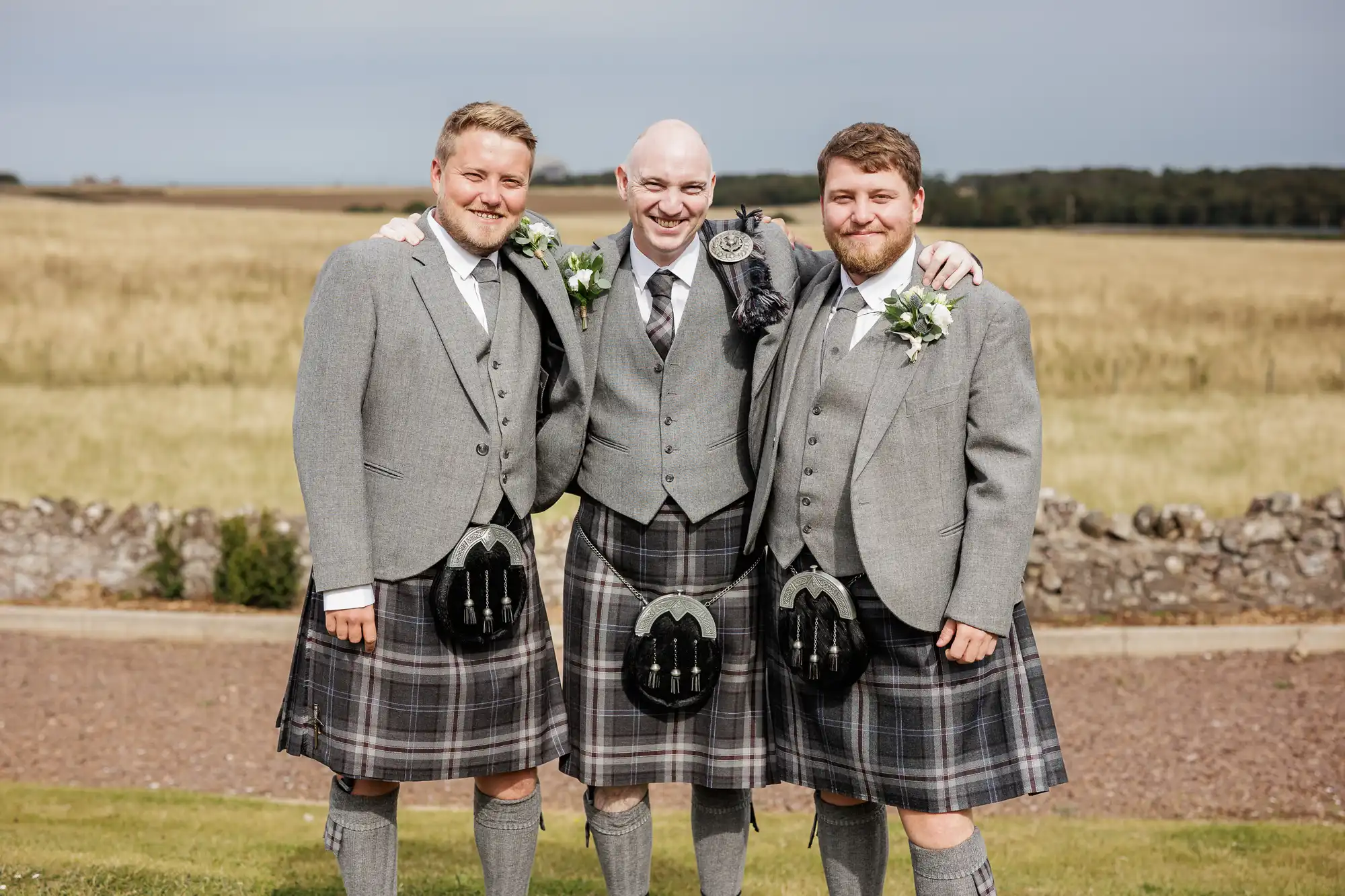 Three men stand outdoors on grass, smiling with arms round each other. All wear grey jackets, tartan kilts, knee-high socks, and sporrans. Two have beards, one is bald. Behind them is a stone wall, fields, and a cloudy sky.