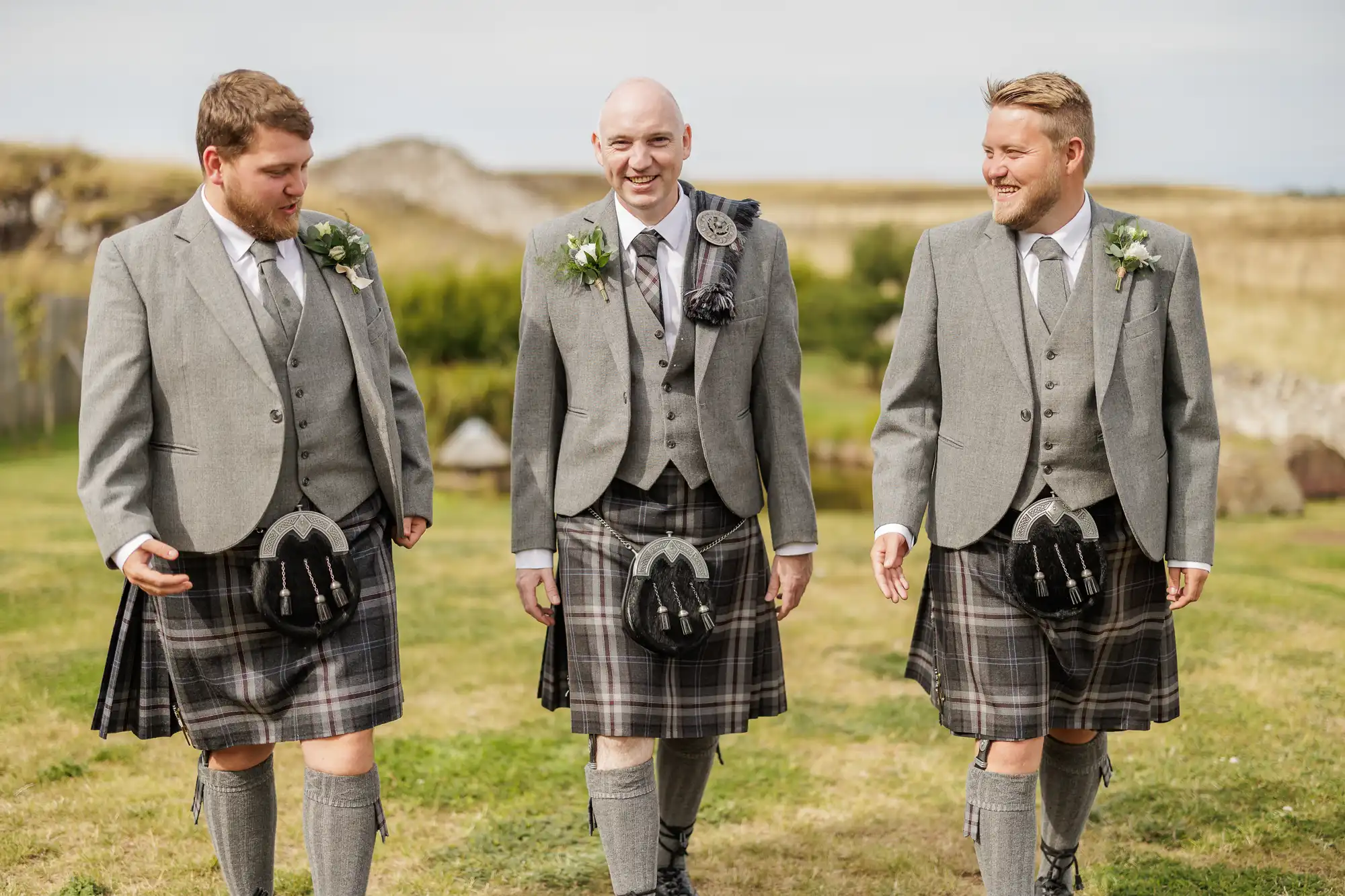 Three men walk outdoors on grass, smiling and wearing traditional Scottish kilts, grey jackets, waistcoats, ties, and sporrans. The background shows fields, low hills, and a partly cloudy sky, suggesting a countryside or rural setting, possibly during a wedding.