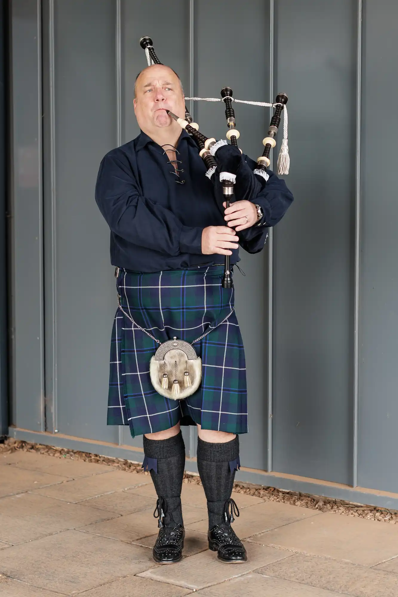A man in a dark blue shirt and traditional blue and green tartan kilt stands on a stone floor, playing bagpipes. He wears long socks, black shoes, and a sporran, with a grey metal wall as the background.