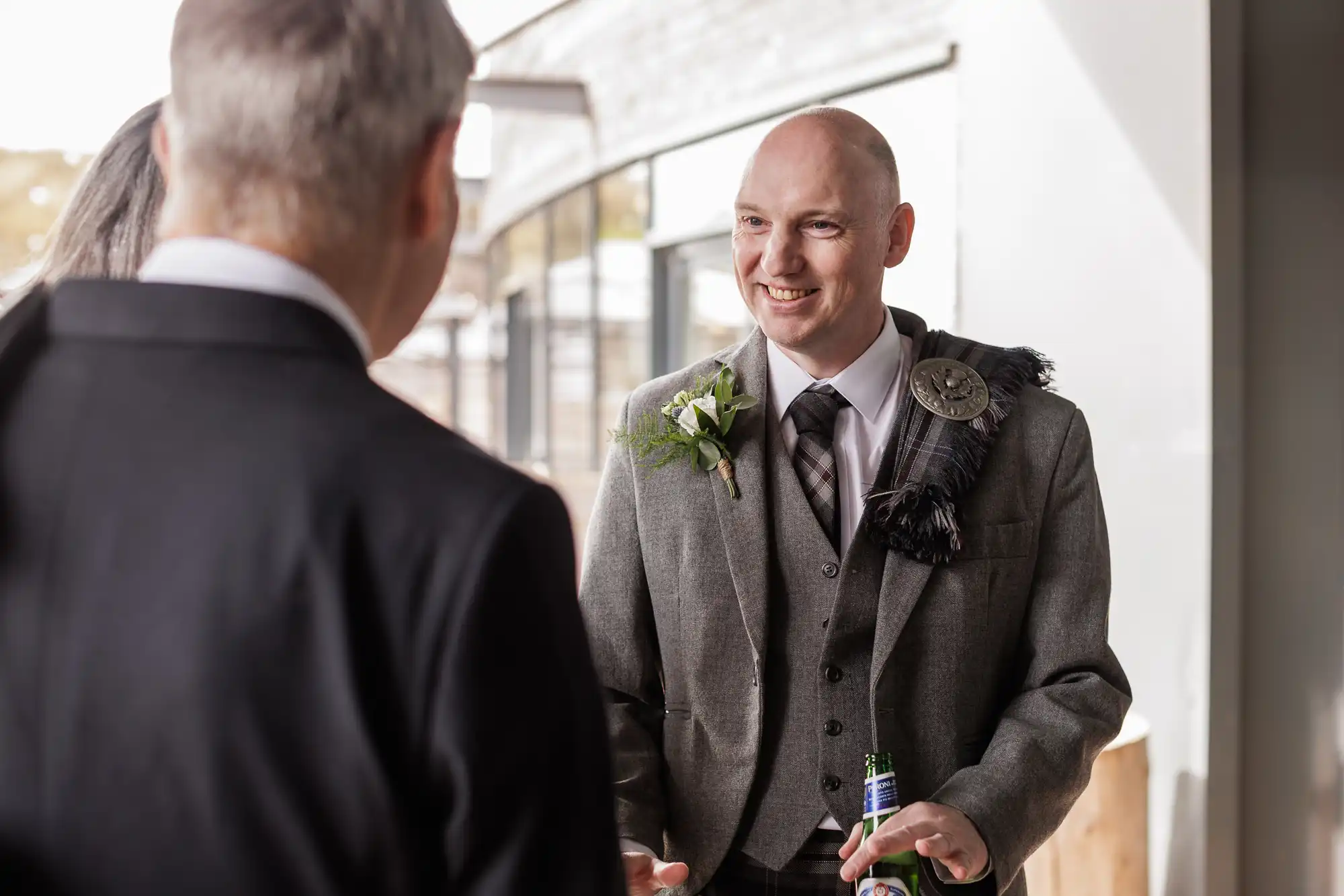 A smiling bald man in a grey suit with a buttonhole and decorative sash speaks with another man in a dark suit. He holds a bottle and stands in a bright indoor space with large windows and natural light.
