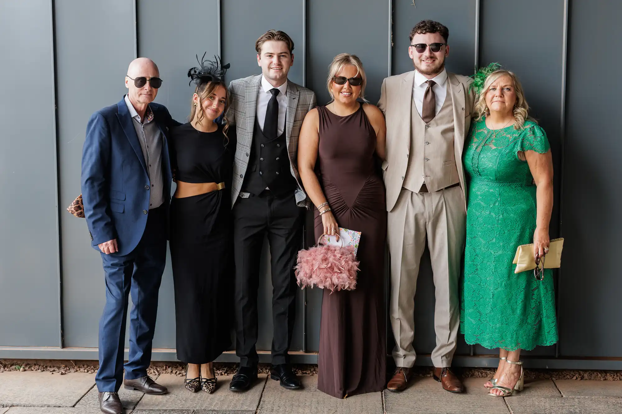 Six adults stand smiling in front of a grey panelled wall. The men wear suits, while the women wear formal dresses in black, brown, and green, with fascinators or accessories. They stand close together, posing for a group photo on a paved surface.