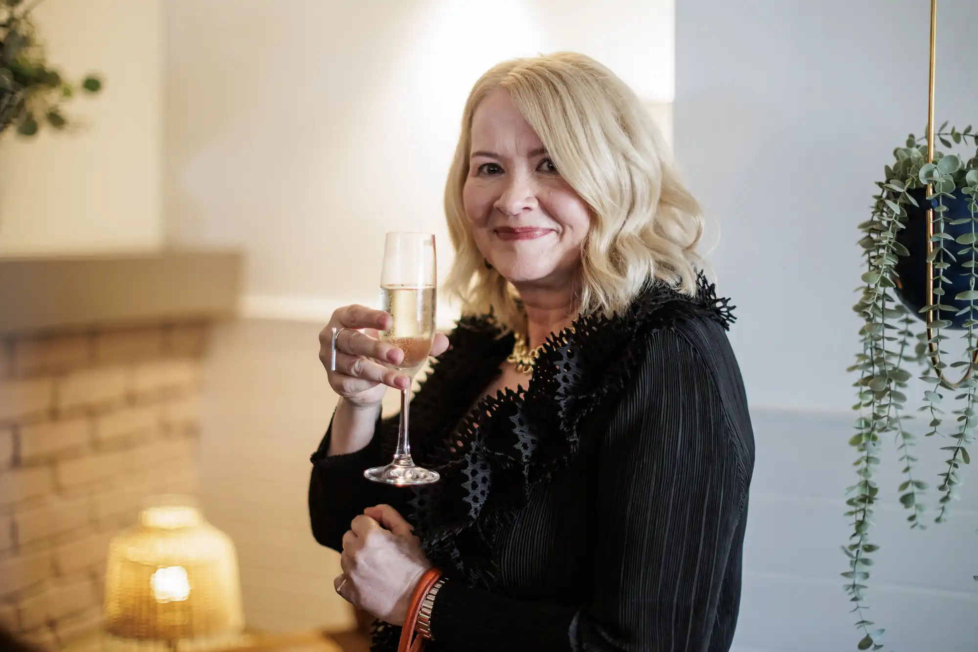 A woman with shoulder-length blonde hair, wearing a black ruffled blouse, smiles and raises a champagne glass. She stands indoors near a white wall decorated with greenery, with warm lighting and a cosy, softly blurred background.