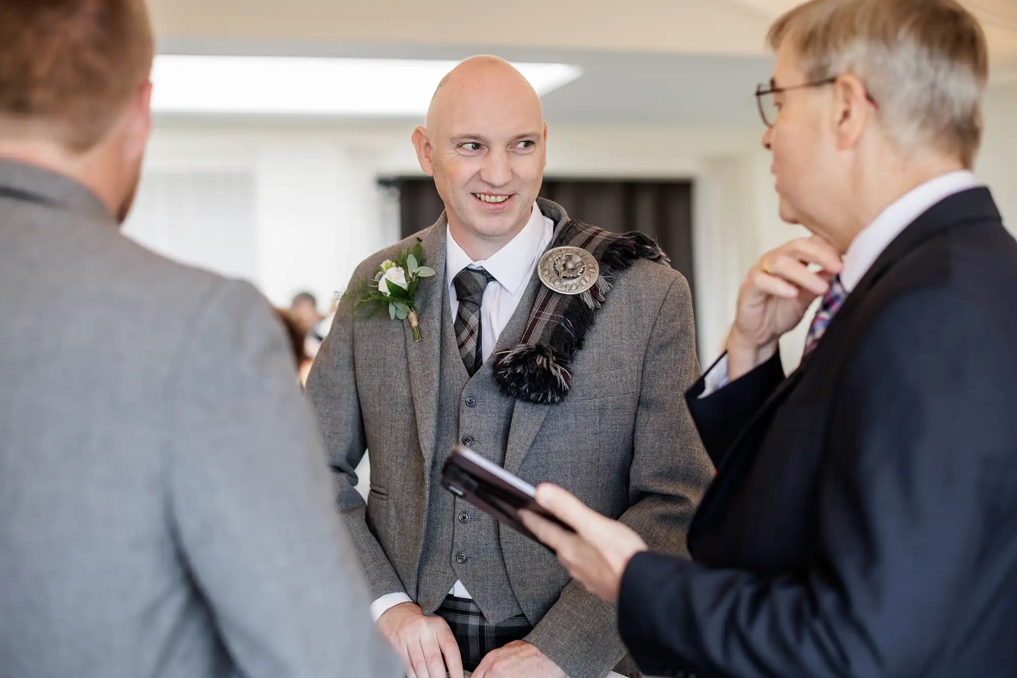 A bald man in a grey Scottish kilt outfit with a buttonhole and sash smiles whilst talking to two men in suits at an indoor event. One man holds a wallet or small notebook; the atmosphere appears formal and friendly.