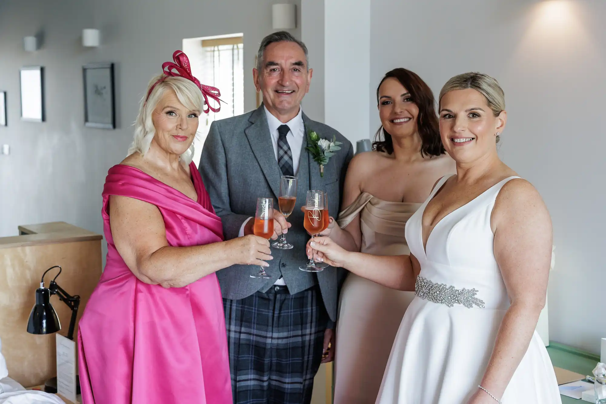 Four adults stand indoors, smiling and holding glasses of pink champagne. The man in the centre wears a grey suit with a kilt; the women wear elegant dresses—one in bright pink with a fascinator, one in cream, and one in a white wedding gown with a jewelled belt.