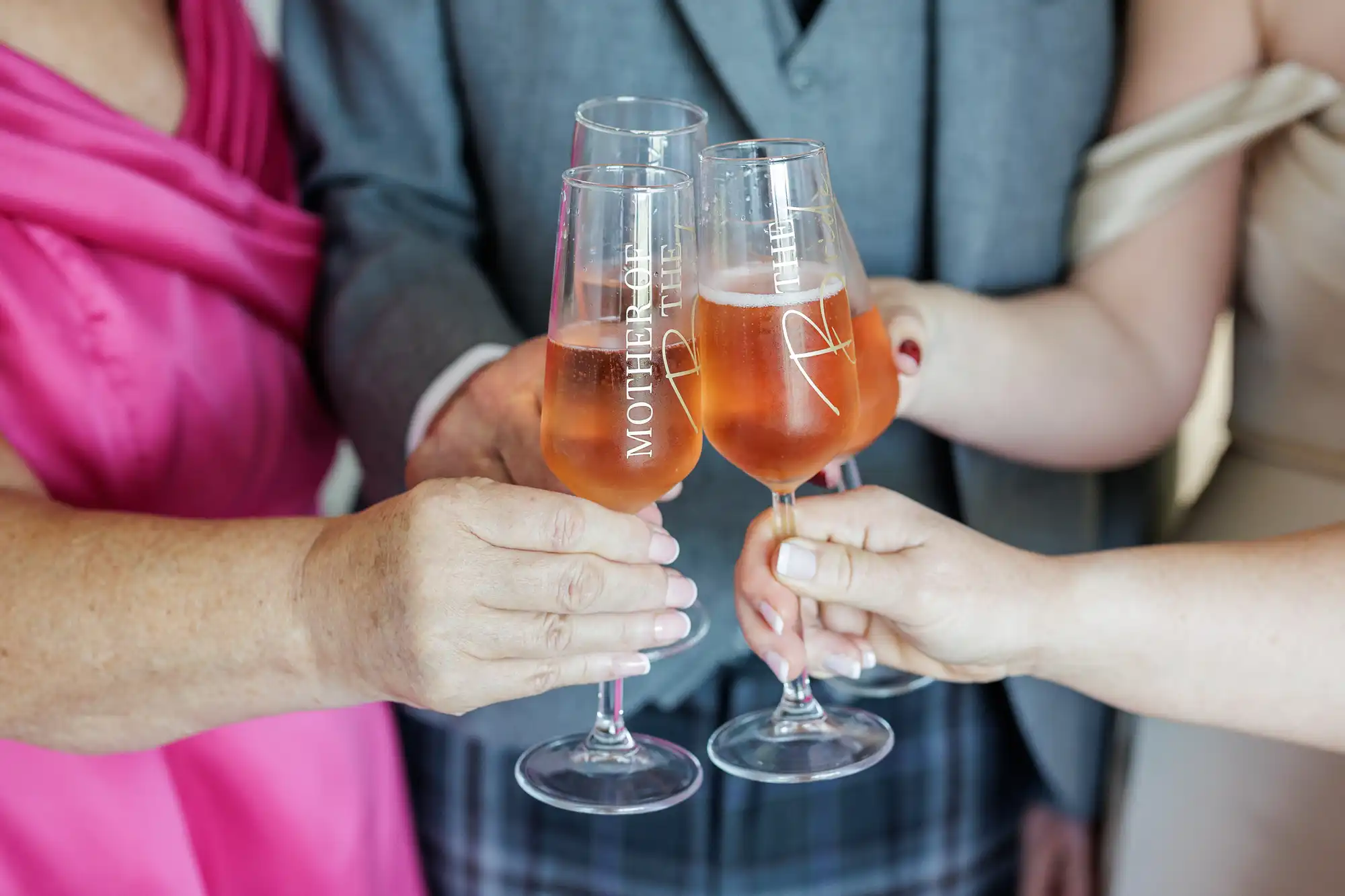 Three people clink glasses of rosé champagne. The glasses are engraved, with one reading “MOTHER OF THE BRIDE.” The people are dressed formally; one wears pink, one is in grey check and a waistcoat, and another wears a pale off-shoulder dress.