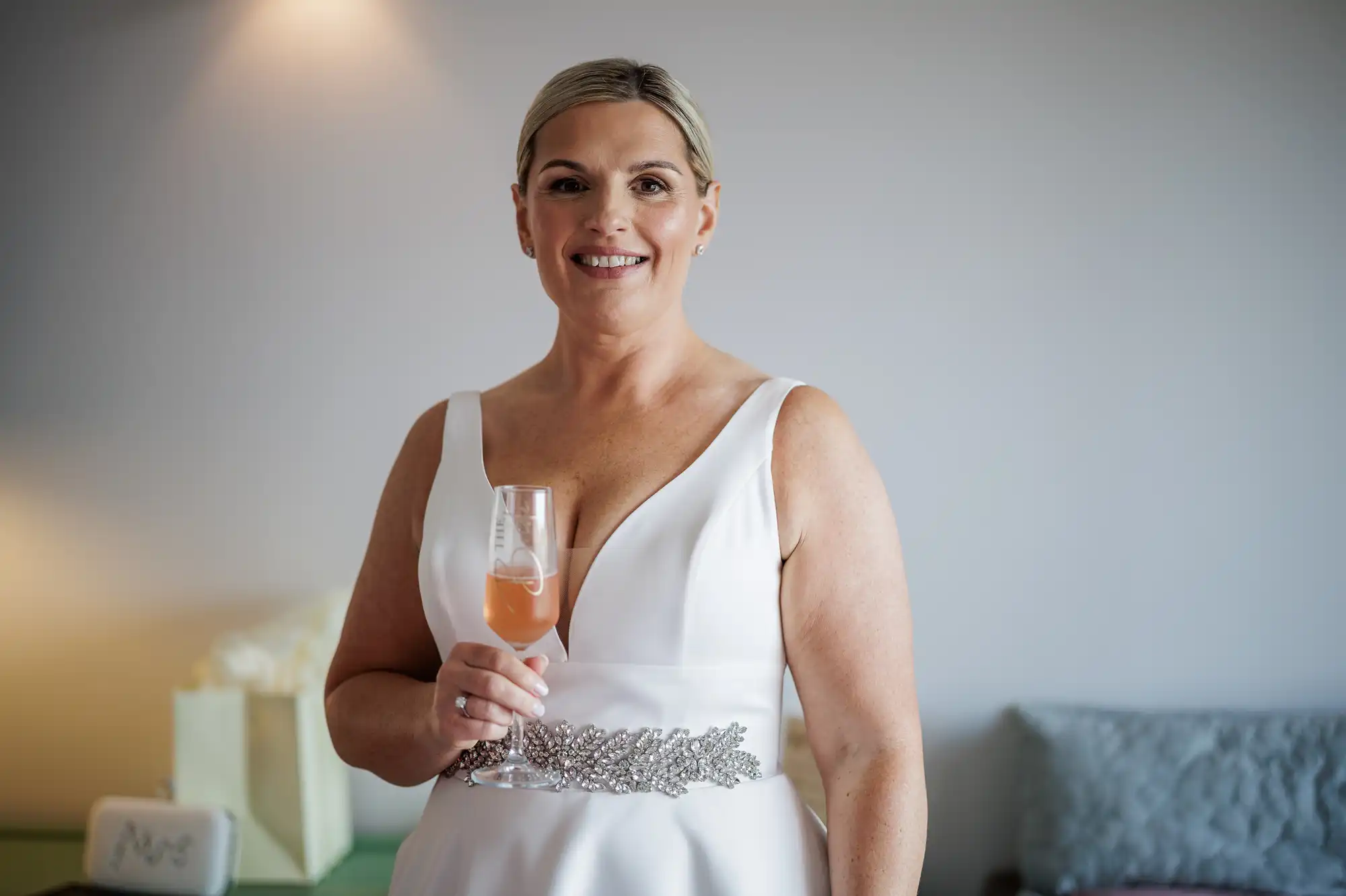 A smiling woman in a white sleeveless dress with a jewelled waistband holds a glass of orange cocktail. She has blonde hair pulled back and stands indoors with soft lighting, a cream gift bag, and grey cushion visible in the background.