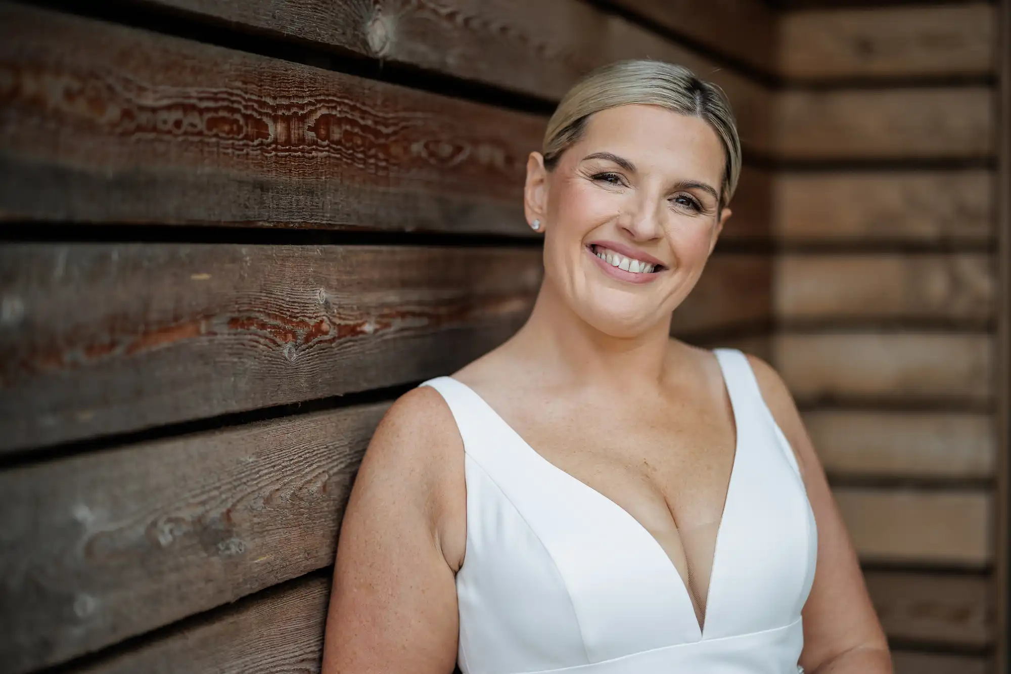 A smiling woman with fair skin and blonde hair, styled back, stands against a wooden wall. She wears a sleeveless white dress with a deep V-neckline and stud earrings, looking directly at the camera in a relaxed pose.