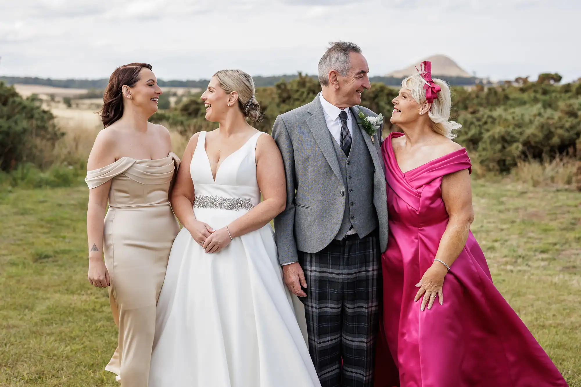 Four people stand outside on grass, smiling at each other. A bride in a white dress stands beside a man in a grey jacket and checked trousers. To their sides are two women: one in a champagne gown, the other in a bright pink dress and matching headband. Shrubs and fields are behind them.