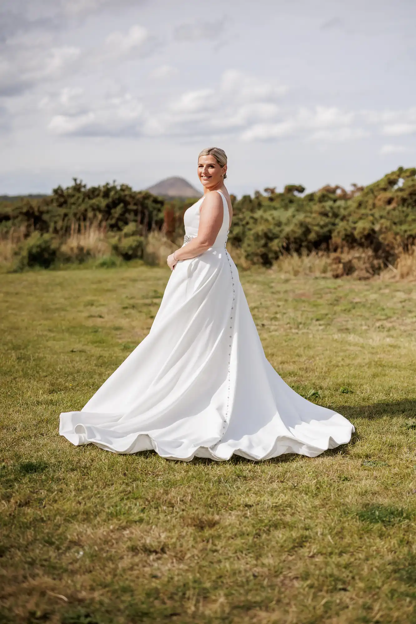 A woman in a sleeveless white wedding dress stands outdoors on grass, smiling and looking over her shoulder. Her gown has a full skirt and button details down the back. Bushes and distant hills are under a partly cloudy sky in the background.
