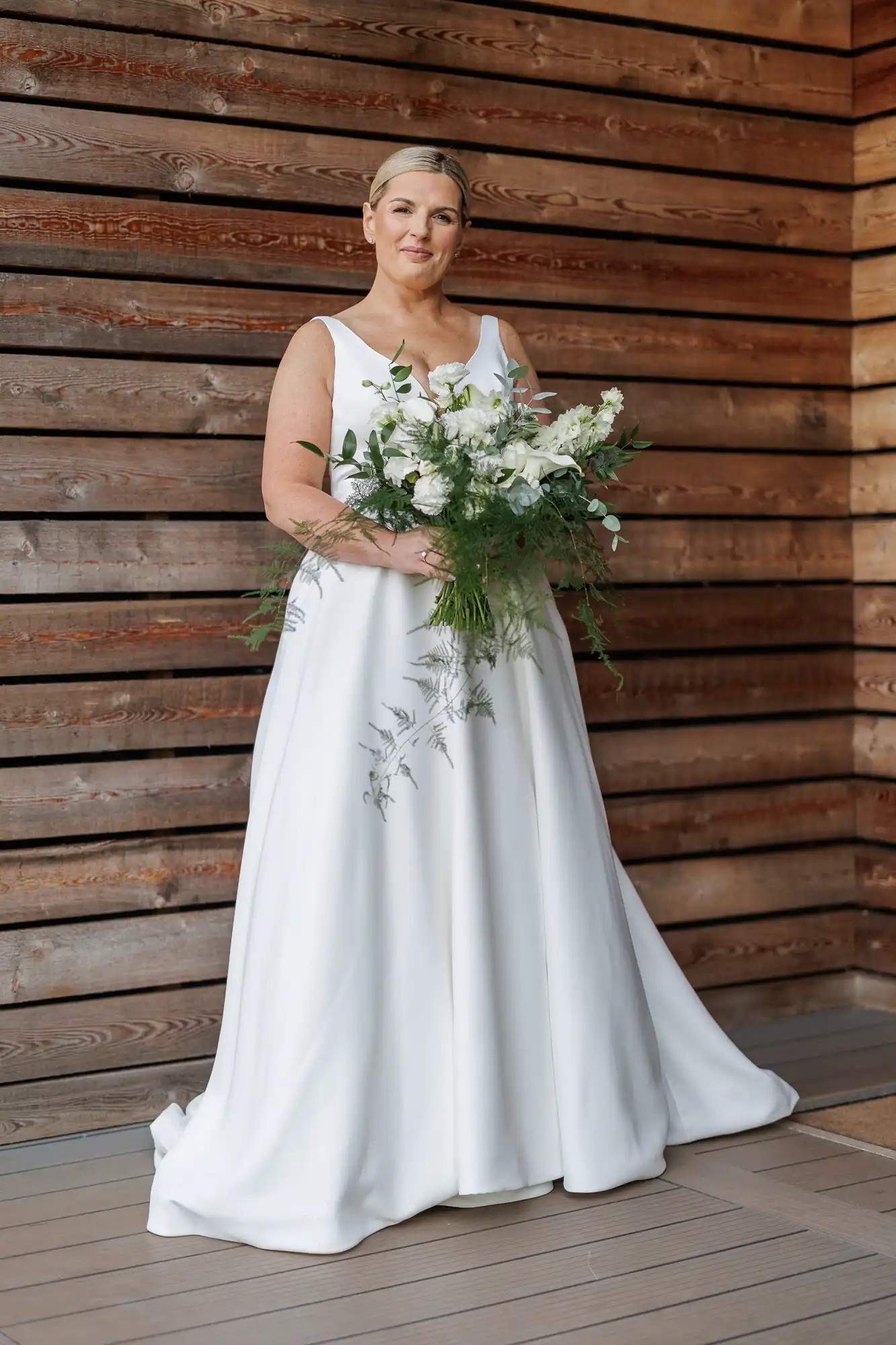 A woman in a sleeveless white wedding dress stands on a wooden decking against a backdrop of horizontal wooden planks, holding a large bouquet of white flowers and greenery. She has short, blonde hair and is smiling gently at the camera.
