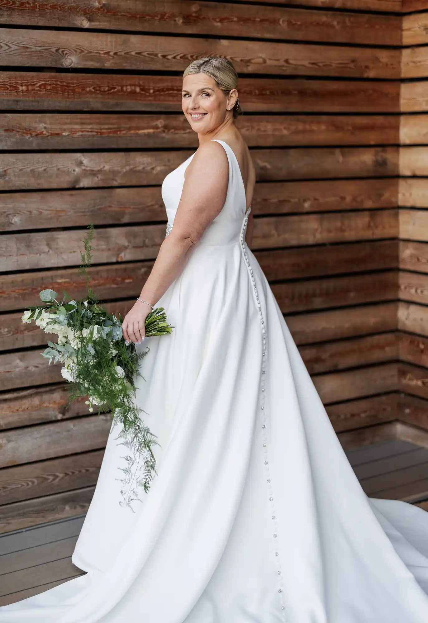 A smiling bride in a white, sleeveless gown with a long train and back buttons holds a green and white bouquet, standing in front of a wooden plank wall, looking over her shoulder. Her blonde hair is styled in a sleek, low bun.