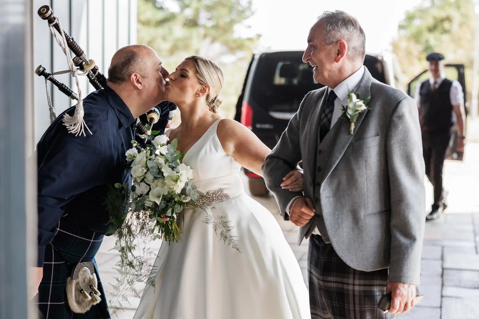A bride in a white dress holding a bouquet kisses a man in a dark shirt and kilt playing bagpipes. An older man in a grey jacket and kilt smiles beside her, linking arms. They are outdoors, with a car and greenery in the background.